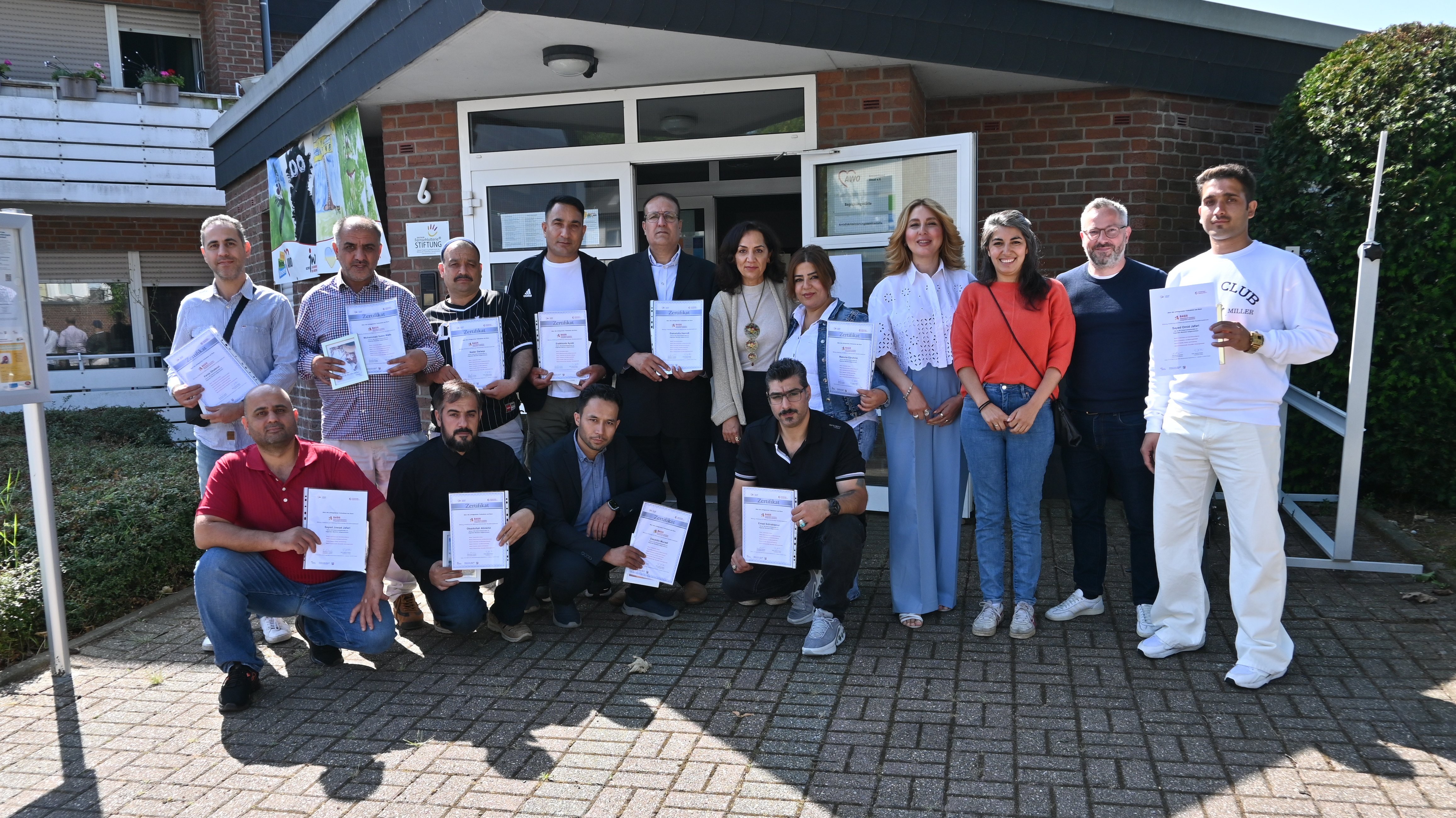 Auf dem Foto ist eine Gruppe von etwa 17 Erwachsenen zu sehen, die draußen vor einem Gebäude posieren. Die meisten von ihnen halten Zertifikate oder Urkunden in ihren Händen. Es scheint eine Art Abschluss- oder Anerkennungsveranstaltung zu sein.