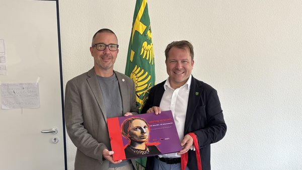Auf dem Foto sind zwei Männer in einem Büro zu sehen, die gemeinsam in die Kamera lächeln. Der Mann links trägt ein Sakko mit grauem T‑Shirt und hält zusammen mit dem Mann rechts ein großes, buntes Plakat oder Buch mit einem Porträt einer Frau darauf. Rechts steht ein Mann im Anzug mit offenem Hemdkragen, der zusätzlich eine rote Stofftragetasche mit weißer Aufschrift (AWO‑Branding) in der Hand hält. Im Hintergrund stehen eine grün-gelbe Fahne bzw. ein Wappenbanner und eine grüne Pflanze, rechts am Bildrand ist ein Drucker auf einem Tisch zu sehen.