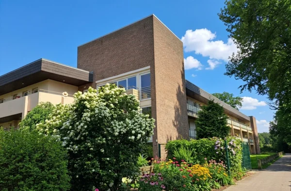Auf dem Bild ist ein mehrstöckiges Wohngebäude mit einer großen Klinkerfassade und seitlichen Balkonen zu sehen. Davor befindet sich ein üppig bepflanzter Garten mit vielen blühenden Sträuchern und Blumen, rechts verläuft ein Weg entlang hoher Bäume.