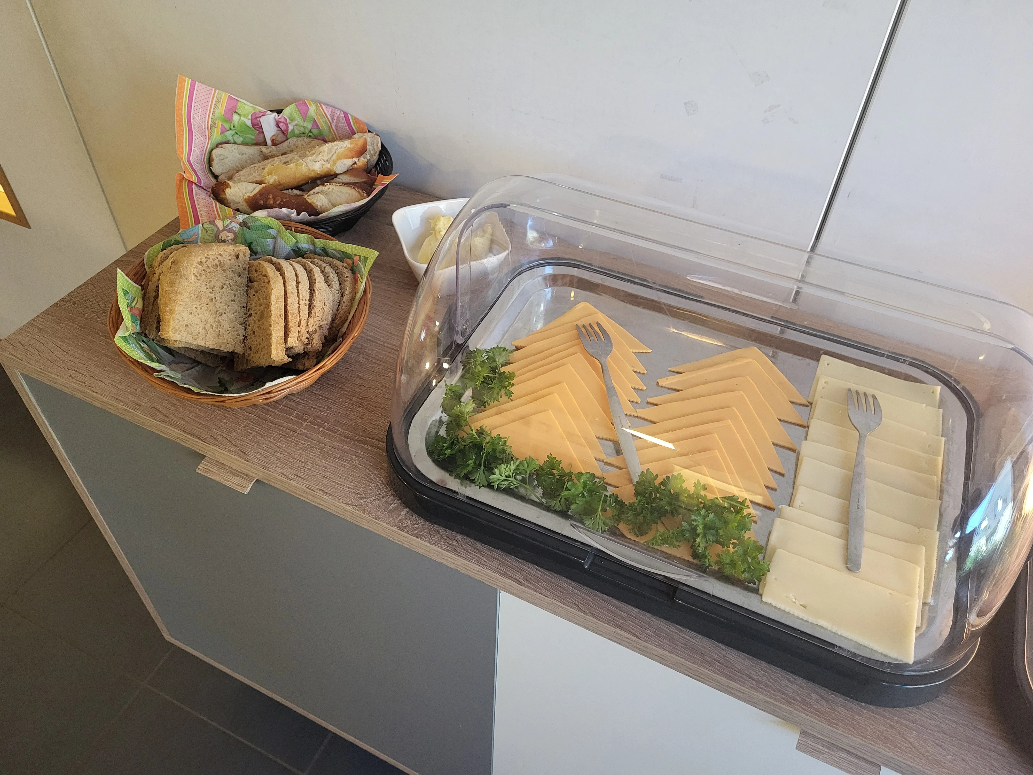 Auf dem Bild ist ein kleines Frühstücksbuffet zu sehen. Zu erkennen sind: Körbe mit geschnittenem Brot und Brötchen Eine große, abgedeckte Platte mit Scheiben von gelbem und weißem Käse Serviergabeln und ein Käsemesser Petersilie als Dekoration Eine Schale mit Butter oder einem weiteren Aufstrich Die Speisen stehen auf einer Anrichte in einem Innenraum bereit.