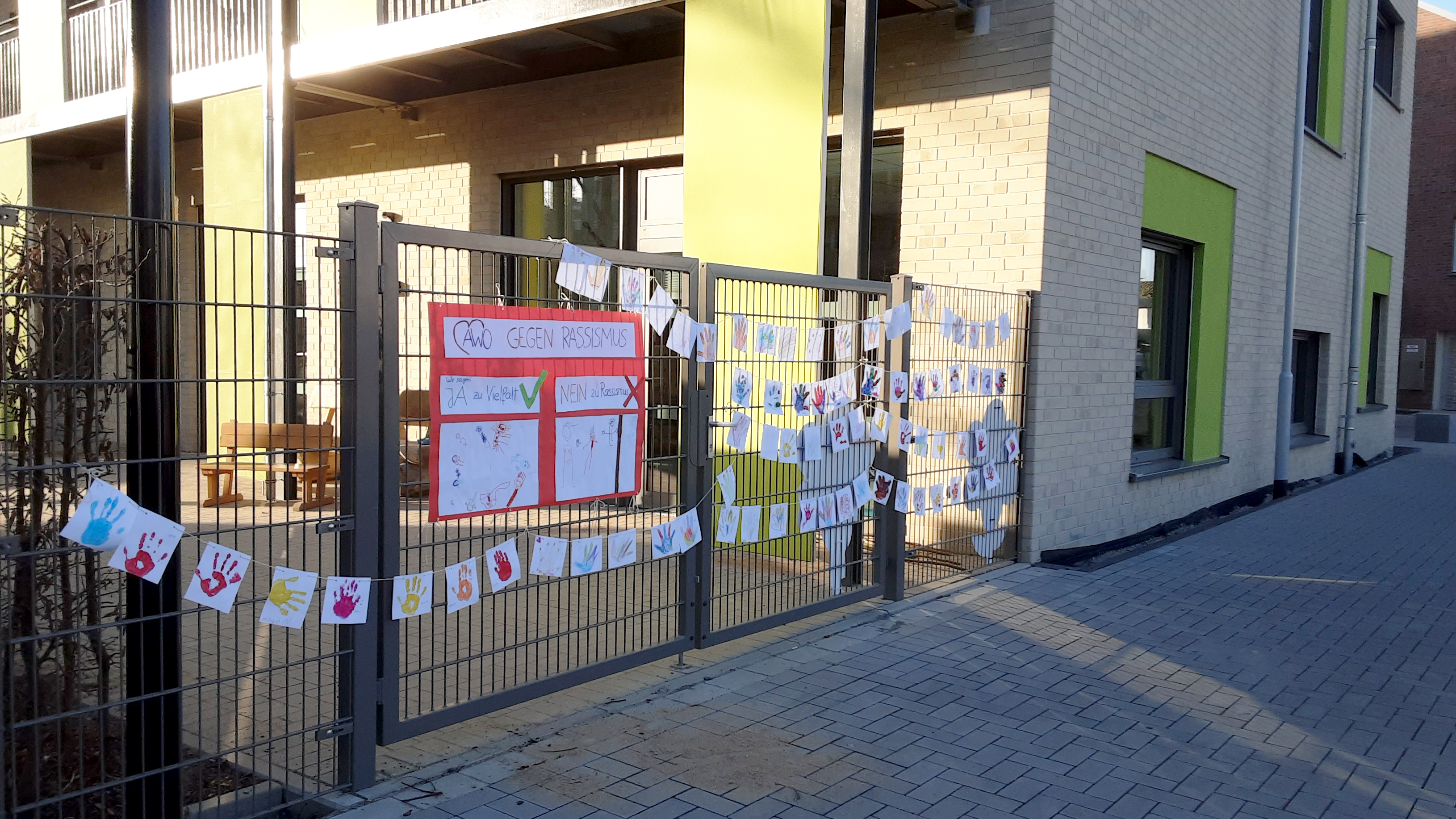 Auf dem Foto ist ein Zaun vor einem modernen Gebäude zu sehen, an dem viele bunte Zeichnungen und Handabdrücke aufgehängt sind. In der Mitte befindet sich ein großes Plakat mit der Aufschrift „Gegen Rassismus“ und den Worten „JA zu Vielfalt“ (mit grünem Haken) und „NEIN zu Rassismus“ (mit rotem Kreuz). Die Zeichnungen scheinen von Kindern gemacht worden zu sein, da sie bunte Handabdrücke und einfache Bilder enthalten. Das Bild vermittelt eine klare Botschaft gegen Rassismus und für Vielfalt, wahrscheinlich im Rahmen eines Schul- oder Gemeinschaftsprojekts. Das Gebäude im Hintergrund sieht aus wie eine Schule oder eine Kindertagesstätte.