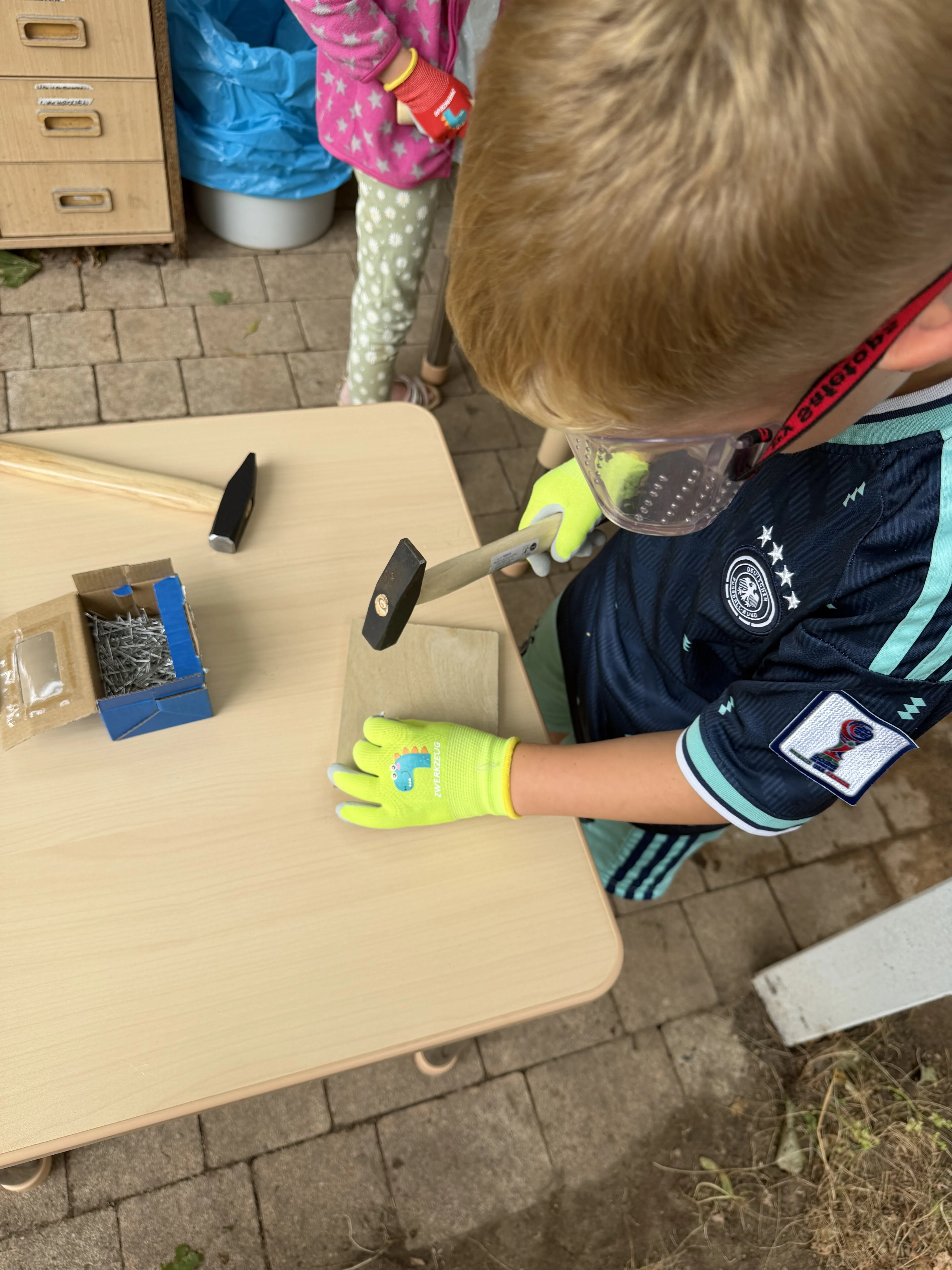 Auf dem Bild arbeitet ein Kind an einem kleinen Holzstück: Es trägt eine Schutzbrille und leuchtend grüne Arbeitshandschuhe. Mit einem Hammer schlägt es vermutlich einen Nagel in das Holz. Auf dem Tisch liegen weitere Hämmer, Holzstäbe und eine Schachtel mit Nägeln. Im Hintergrund ist ein weiteres Kind beziehungsweise eine Person mit einer pink-grünen Kleidung zu sehen.