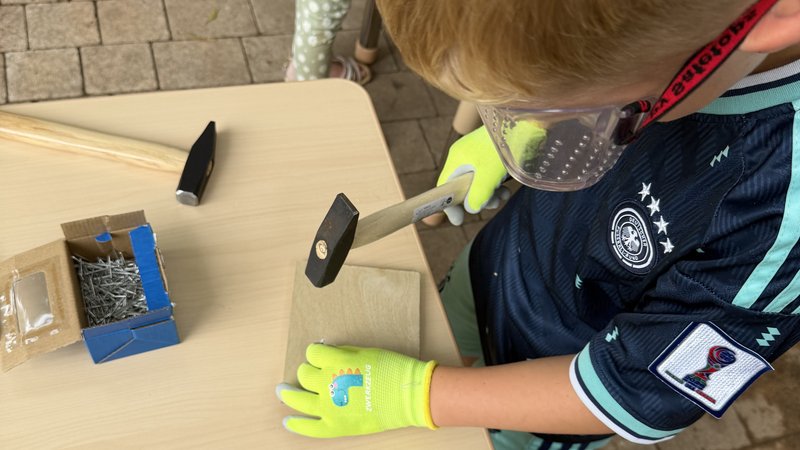 Auf dem Bild arbeitet ein Kind an einem kleinen Holzstück: Es trägt eine Schutzbrille und leuchtend grüne Arbeitshandschuhe. Mit einem Hammer schlägt es vermutlich einen Nagel in das Holz. Auf dem Tisch liegen weitere Hämmer, Holzstäbe und eine Schachtel mit Nägeln. Im Hintergrund ist ein weiteres Kind beziehungsweise eine Person mit einer pink-grünen Kleidung zu sehen.