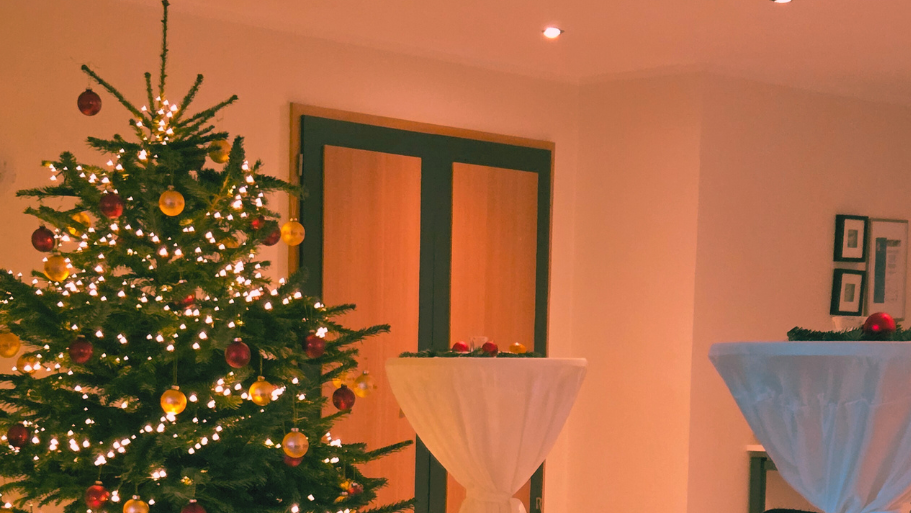 Das Bild zeigt eine festliche, weihnachtliche Szene, wahrscheinlich von einer Jahresabschlussfeier oder einem ähnlichen Anlass, wie der Dateiname "Jahresabschlussfeier 2025.jpg" andeutet. Hier sind die Hauptbestandteile, die zu sehen sind: ? Im Hintergrund steht ein großer, geschmückter Weihnachtsbaum mit Lichtern und goldenen sowie roten Kugeln. ?️ Im Vordergrund ist ein gedeckter Tisch oder eine Tafel, bedeckt mit einem dunkelroten Tuch. Darauf stehen Geschirr (weiße Tassen und Untertassen), Gläser und eine Glasschüssel. Es gibt weihnachtliche Dekoration und Essen, wie Tannenzweige, Orangen/Mandarinen und Erdnüsse (oder ähnliche Nüsse) auf dem Tisch. Links unten ist ein verpacktes Geschenk mit einer roten Schleife zu sehen. ? Im Mittelgrund/Hintergrund stehen zwei hohe, weiß abgedeckte Stehtische (wahrscheinlich für ein Buffet oder Getränke). ⚫ Zwei schwarze Stuhllehnen sind links und rechts im Vordergrund erkennbar. ? Die Beleuchtung ist warm und gedämpft, was eine gemütliche Atmosphäre schafft.