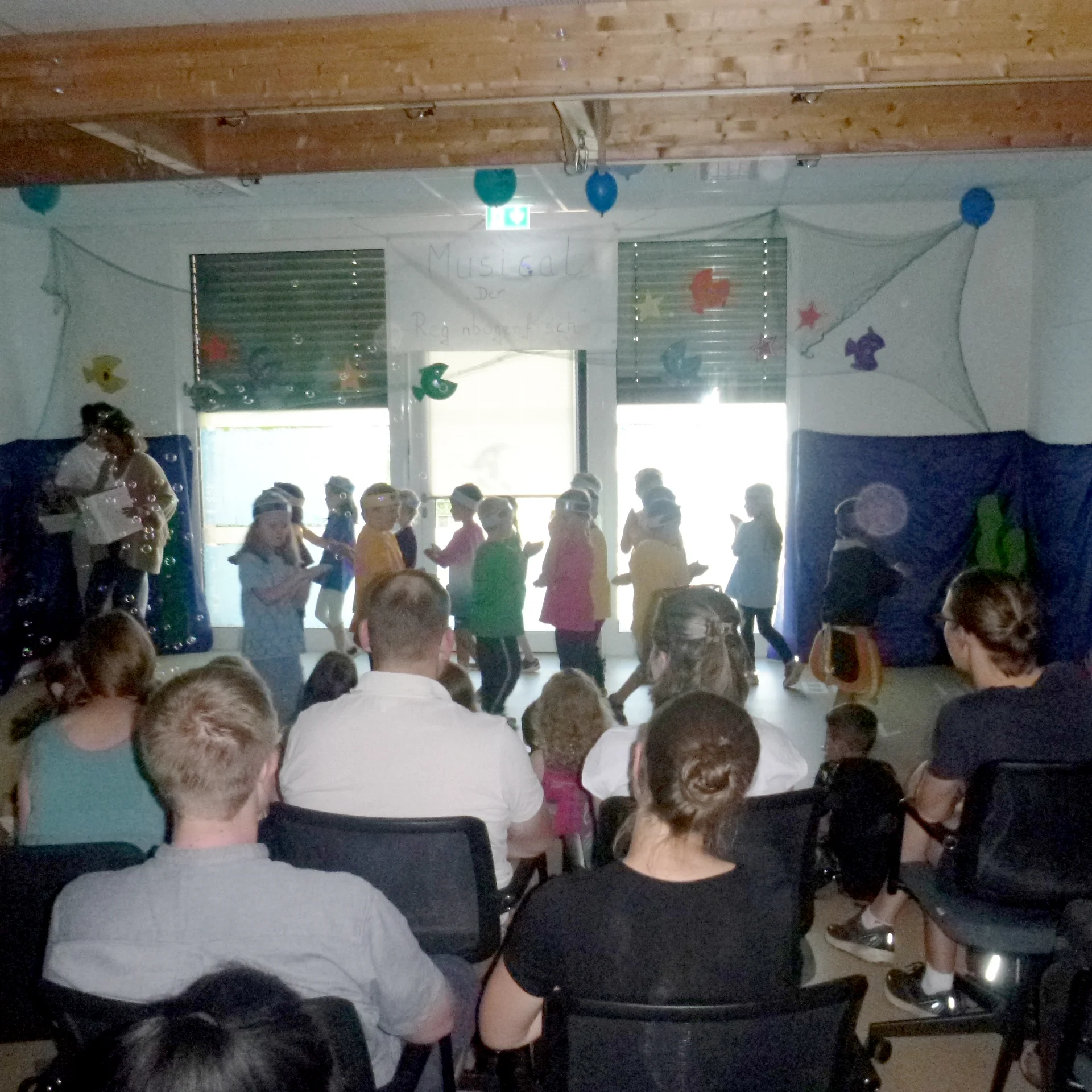 Das Foto zeigt eine Szene in einem Innenraum, in dem sich eine Gruppe von Kindern vor einem Raum versammelt hat. Sie führen eine Aktivität durch. Erwachsene sitzen und beobachten die Kinder. Der Raum ist mit Luftballons und anderen Dekorationen geschmückt, was darauf hindeutet, dass es sich um ein besonderes Ereignis oder eine Feier handelt.