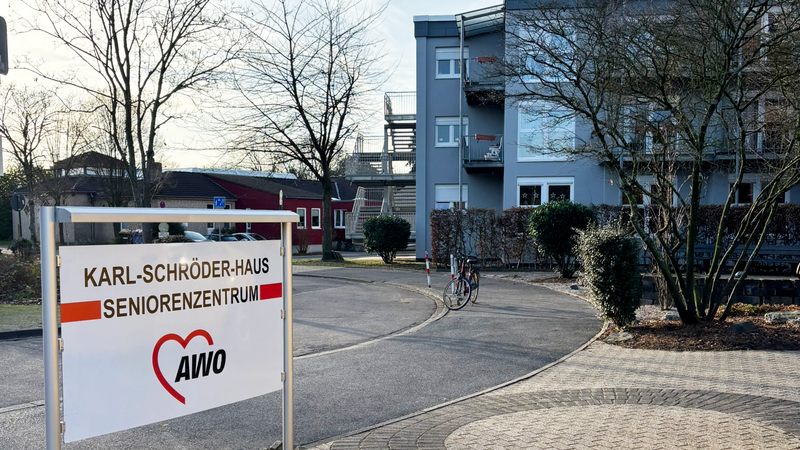 Auf dem Bild ist der Eingangsbereich eines Seniorenzentrums der AWO zu sehen. ​ Beschriftung und Gebäude Im Vordergrund steht ein Schild mit der Aufschrift „Karl-Schröder-Haus Seniorenzentrum“ und dem AWO-Herzlogo. ​ Dahinter sieht man das mehrstöckige Hauptgebäude mit Balkonen sowie weitere flache Gebäude auf dem Gelände. ​ Umgebung Links und rechts des Weges stehen Bäume und Büsche, viele davon ohne Laub, was auf die kalte Jahreszeit hinweist. ​