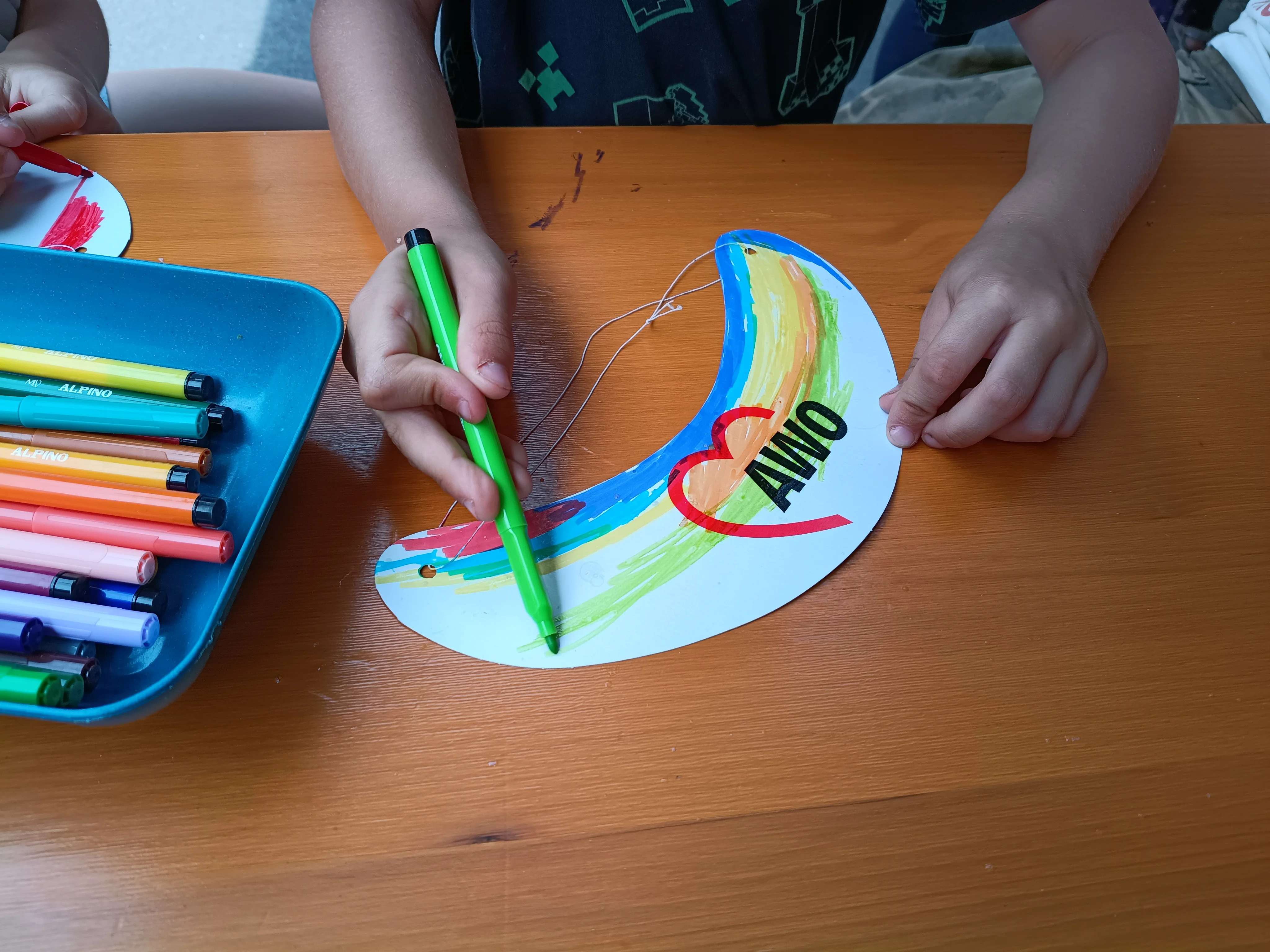 Auf dem Foto sieht man Kinderhände, die an einem Holztisch ein kleines, halbkreisförmiges Pappschild mit dem AWO‑Logo bunt ausmalen. Links steht ein Tablett mit verschiedenen farbigen Filzstiften, rechts hält ein Kind einen grünen Stift und gestaltet damit den Regenbogen auf dem Schild weiter.