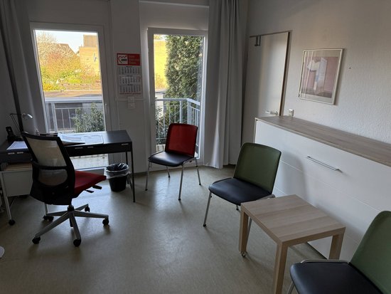 Das Bild zeigt ein Zimmer mit mehreren Möbelstücken. Links im Bild steht ein Schreibtisch mit einem schwarzen Bürostuhl davor. Auf dem Schreibtisch befinden sich einige Gegenstände, darunter ein Computerbildschirm und eine Lampe. Neben dem Schreibtisch steht ein kleiner schwarzer Mülleimer. In der Mitte des Raumes stehen drei Stühle: einer mit rotem Sitzpolster, einer mit grünem Sitzpolster und einer mit schwarzem Sitzpolster. Vor den Stühlen befindet sich ein kleiner Holztisch. Rechts im Bild ist eine Kommode zu sehen, auf der eine gerahmte Zeichnung oder ein Bild hängt. Im Hintergrund sind zwei Fenster zu sehen, die viel Licht in den Raum lassen und einen Blick nach draußen ermöglichen.