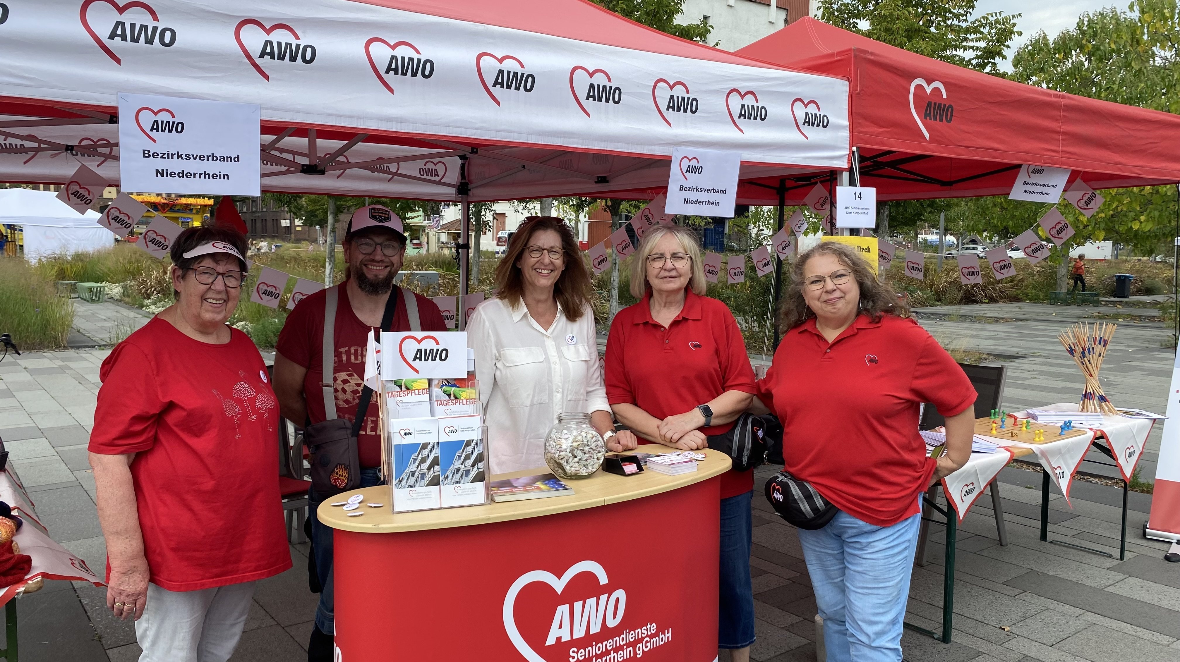 Auf dem Foto sind fünf Personen an einem Stand der AWO (Arbeiterwohlfahrt) zu sehen. Der Stand besteht aus zwei roten Pavillons, die mit dem AWO-Logo und Herzen bedruckt sind. Auf den Schildern am Stand steht "AWO Bezirksverband Niederrhein". Die Personen tragen AWO-T-Shirts und stehen hinter einem kleinen Tisch, auf dem Informationsmaterial ausliegt. Im Hintergrund sind weitere Stände und Bäume zu erkennen. Es handelt sich wahrscheinlich um eine Veranstaltung im Freien.