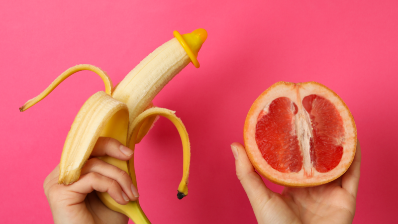 Auf dem Foto sind zwei Hände zu sehen, die einen Bananen- und einen Grapefruit-Kondom-Ständer halten. Die Banane ist teilweise geschält und trägt einen übergestülpten Kondom. Die Grapefruit ist in der Mitte halbiert und dient als Metapher für weibliche Geschlechtsorgane.
