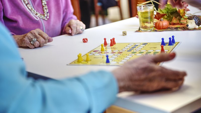 Auf dem Foto sind mehrere ältere Personen zu sehen, die gemeinsam ein Brettspiel spielen. Es handelt sich um das Spiel „Mensch ärgere dich nicht“. Auf dem Tisch liegt das Spielbrett mit bunten Spielfiguren (gelb, blau, rot) und zwei Würfeln. Die Szene spielt sich vermutlich in einem Gemeinschaftsraum ab – vielleicht in einem Seniorenheim oder einer Betreuungseinrichtung. Im Hintergrund steht ein Glas mit einem Getränk und herbstlicher Dekoration (z. B. kleine Kürbisse und ein Igel). Die Kleidung und der Schmuck der Personen sowie die liebevolle Tischdekoration lassen eine warme, gepflegte Atmosphäre erkennen. Das Bild vermittelt Gemeinschaft, Aktivität und Lebensfreude im Alter.