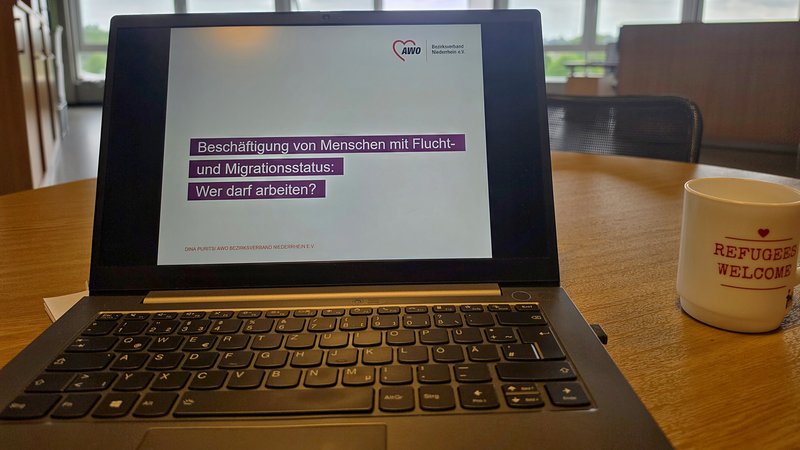 Auf dem Foto ist ein aufgeklappter Laptop zu sehen, auf dessen Bildschirm eine PowerPoint-Folie mit dem AWO-Logo und dem Titel "Beschäftigung von Menschen mit Flucht- und Migrationsstatus: Wer darf arbeiten?" von Dina Purits vom AWO Bezirksverband Niederrhein e.V. angezeigt wird. Der Laptop steht auf einem Holztisch in einem Büro- oder Konferenzraum mit großen Fensterfronten, durch die ein bewölkter Himmel und Grünflächen zu erkennen sind. Rechts daneben steht eine Kaffeetasse mit der Aufschrift "REFUGEES WELCOME" und einem Herz-Symbol