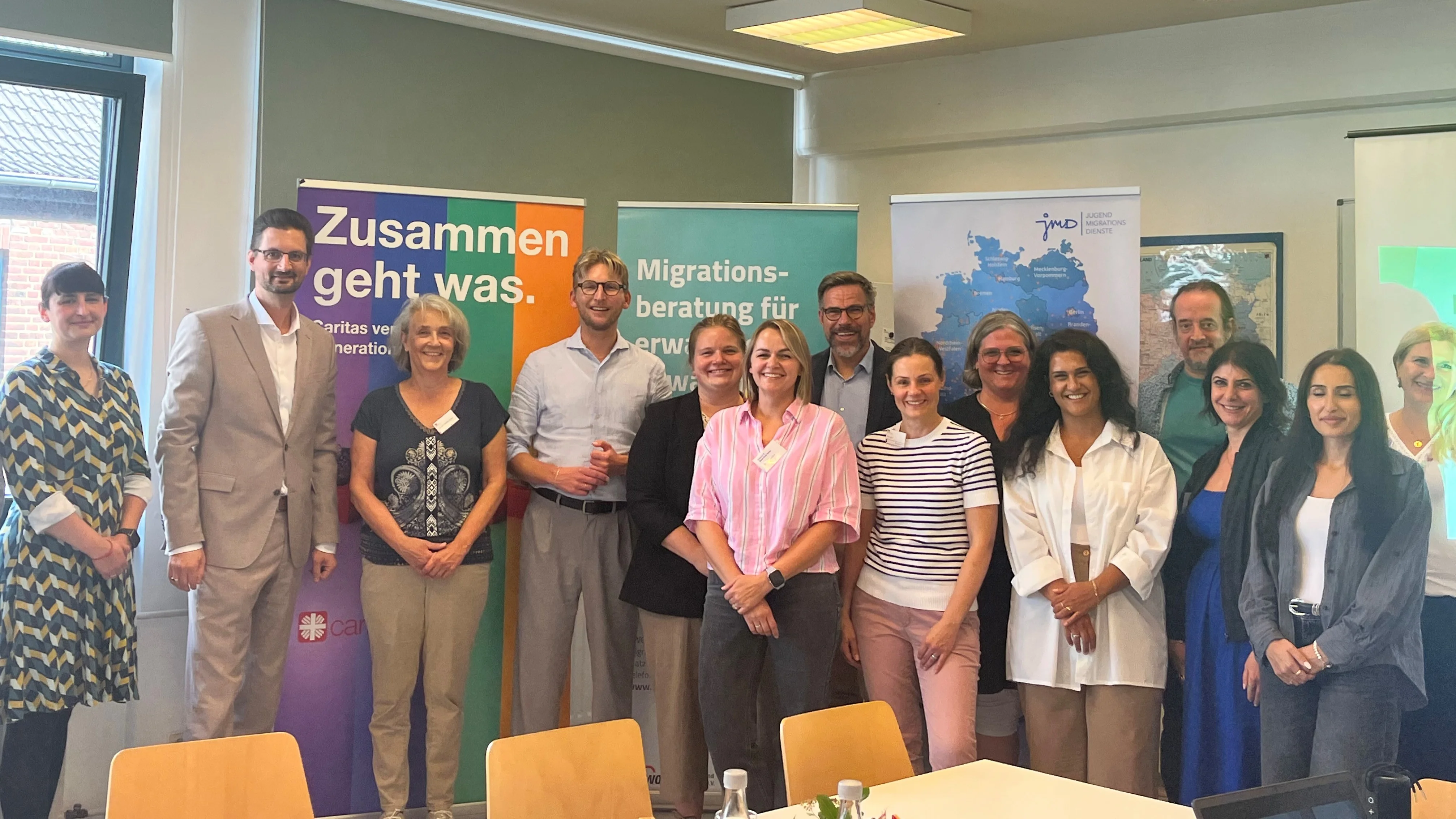 Auf dem Bild ist eine Gruppe von etwa 14 Personen in einem Besprechungsraum zu sehen. Sie stehen für ein gemeinsames Gruppenfoto vor mehreren Roll-up-Bannern mit Hinweisen auf Migrationsberatung, Jugendmigrationsdienste und die Caritas. Im Hintergrund befinden sich außerdem Fenster mit heruntergelassenen Jalousien, Deckenleuchten, eine Deutschlandkarte sowie ein Flipchart. Im Vordergrund sind Tische, Stühle und Getränkeflaschen zu sehen.