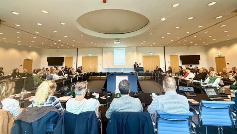 Auf dem Foto ist eine Konferenz oder ein Meeting in einem großen Raum zu sehen. Hier sind die Details: Veranstaltung: Es scheint eine geschäftliche oder akademische Versammlung zu sein, bei der viele Personen anwesend sind. Teilnehmer: Eine große Gruppe von Menschen sitzt an Tischen, die in einem U- oder Rechteckmuster angeordnet sind. Die meisten Gesichter sind von hinten oder von der Seite zu sehen. Raumgestaltung: Der Raum hat eine helle Decke mit einer runden Aussparung in der Mitte. Es gibt mehrere Türen im Hintergrund und zwei große Flachbildschirme an den Seiten. Vortrag/Präsentation: In der Mitte des "U" steht ein Rednerpult oder Tisch, von dem aus eine Person zu sprechen scheint. Eine Leinwand oder ein großer Projektionsbereich im Hintergrund zeigt eine Präsentation (Text oder Grafiken sind nicht klar lesbar). Zusammenfassend handelt es sich um eine Innenaufnahme einer Versammlung oder Tagung mit einem Redner.