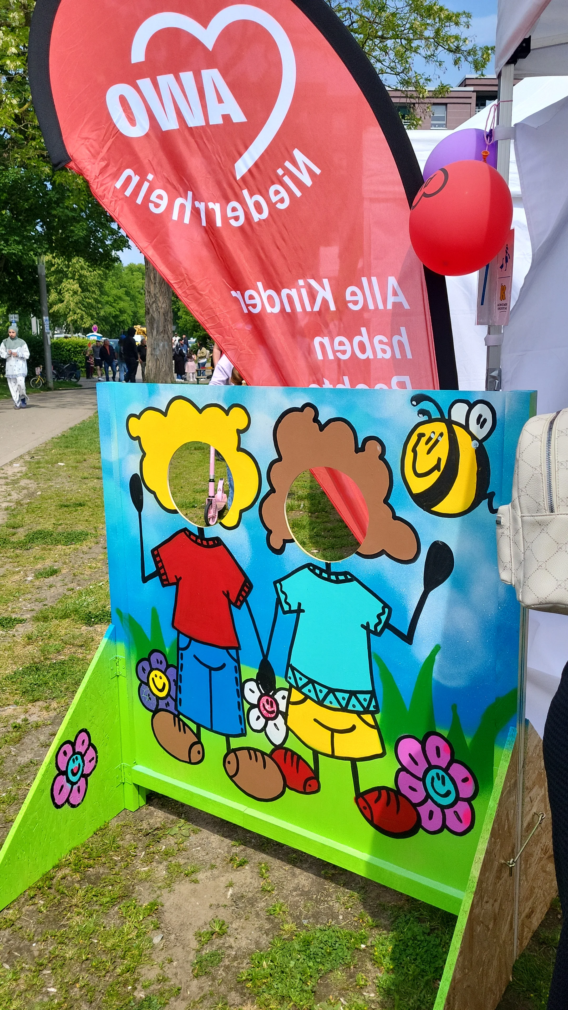 Auf dem Bild ist ein bunter Mitmach‑Aufsteller für Kinder zu sehen: eine bemalte Tafelkonstruktion mit zwei gezeichneten Kinderfiguren, bei denen die Gesichter als runde Löcher ausgespart sind, damit Kinder dort ihre Köpfe hindurchstecken und sich fotografieren lassen können. Daneben steht ein roter AWO‑Niederrhein‑Beachflag‑Banner, außerdem sind Luftballons und Teile eines weißen Pavillons zu erkennen