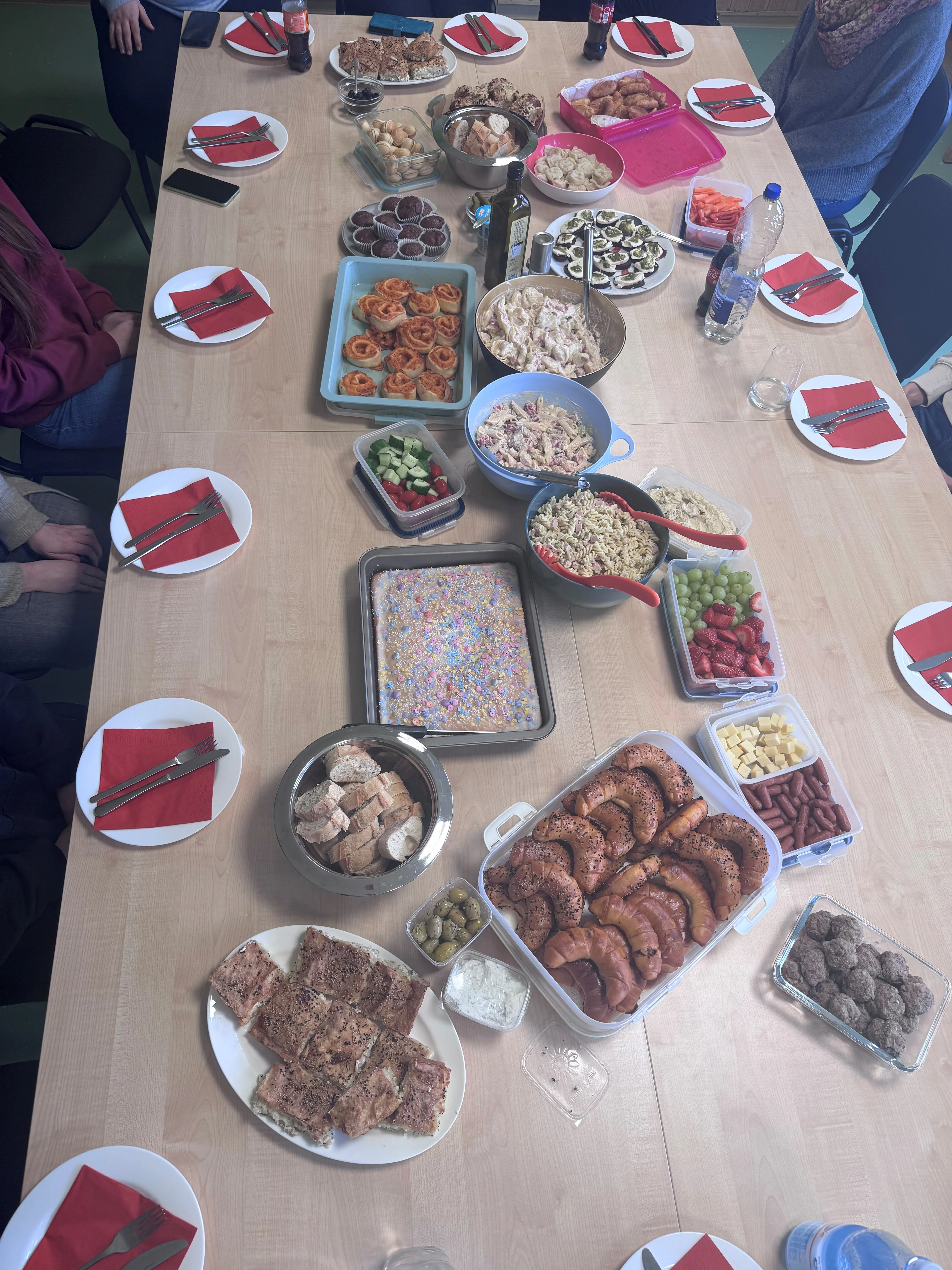 Auf dem Bild ist eine lange, rechteckige Tafel mit einem Buffet aus vielen mitgebrachten Speisen zu sehen. Rund um den Tisch sitzen mehrere Personen, vor denen bereits Teller mit roten Servietten und Besteck bereitliegen.
