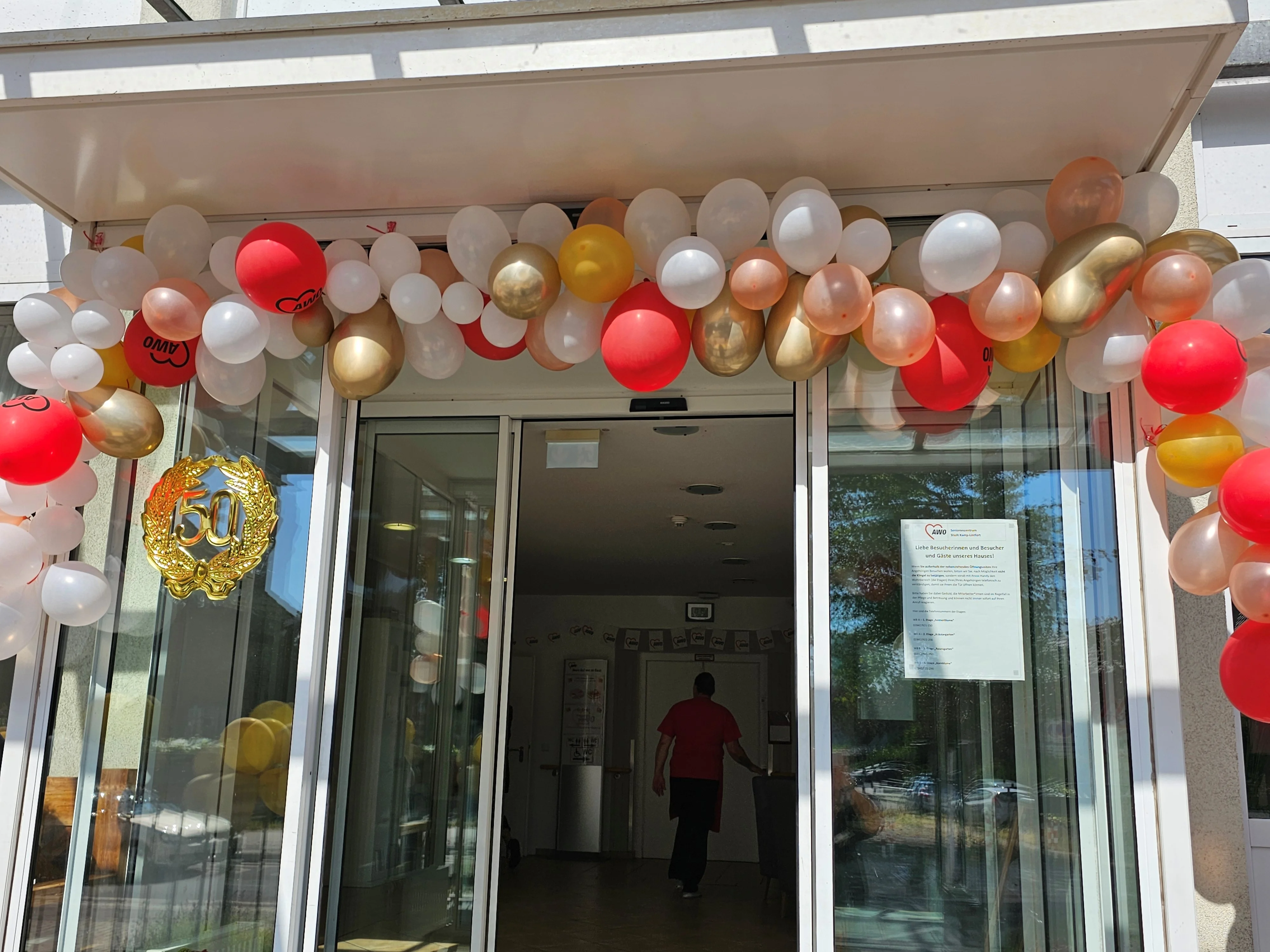 Auf dem Bild ist ein festlich geschmückter Eingangsbereich eines Gebäudes zu sehen. Über der Eingangstür hängt ein dichter Bogen aus Luftballons in Weiß, Rot, Gold und Champagner‑Tönen, einige rote Ballons tragen das AWO‑Logo. In der Mitte der Glasfront klebt ein goldener Kranz mit der Zahl „50“, der auf ein 50‑jähriges Jubiläum hinweist. Hinter der automatischen Schiebetür erkennt man einen Flur, in dem eine Person in roter Oberbekleidung ins Gebäudeinnere geht. Auf der rechten Glastür ist außerdem ein ausgedruckter Hinweistext der Einrichtung angebracht