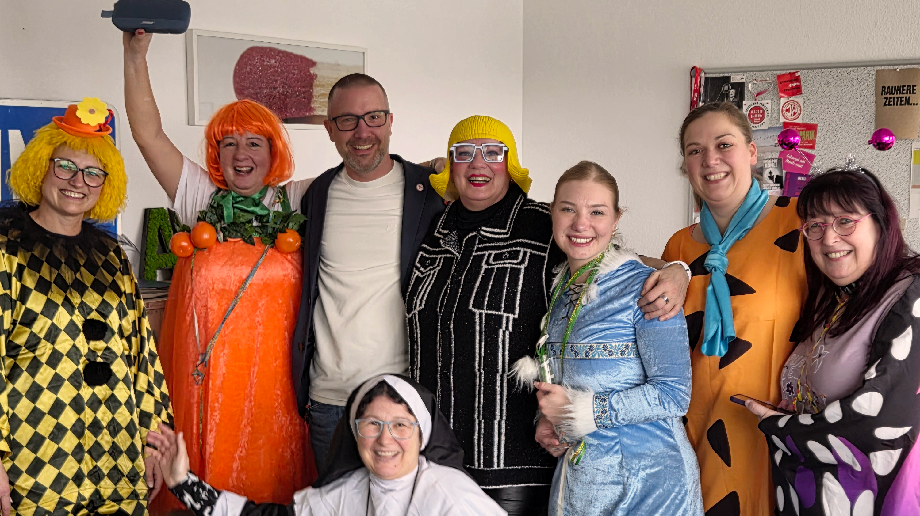 Auf dem Bild ist eine Gruppe von Menschen in bunten Karnevalskostümen in einem Büro‑ bzw. Besprechungsraum zu sehen. Einige tragen auffällige Verkleidungen und alle stehen bzw. knien eng beieinander und posieren fröhlich für das Foto.