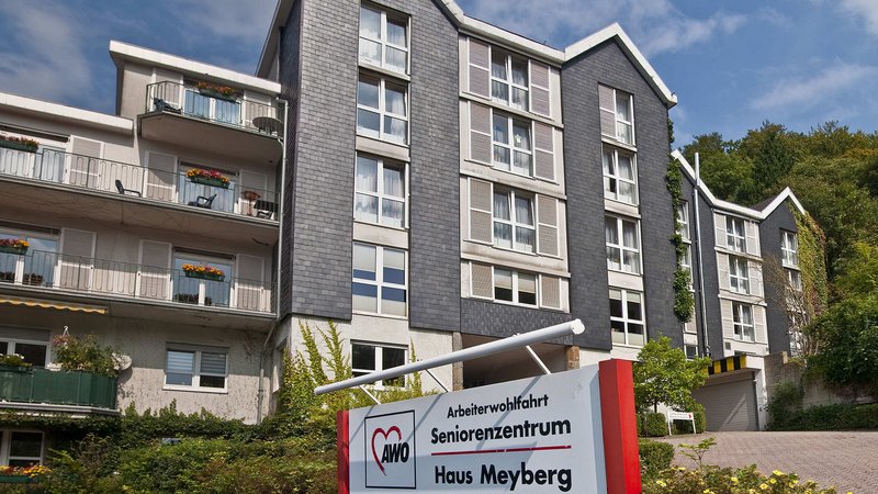 Auf dem Bild ist ein mehrstöckiges Gebäude mit Balkonen zu sehen, offenbar ein Wohn- oder Seniorenzentrum. Im Vordergrund steht ein Schild mit der Aufschrift „Arbeiterwohlfahrt Seniorenzentrum Haus Meyberg“.