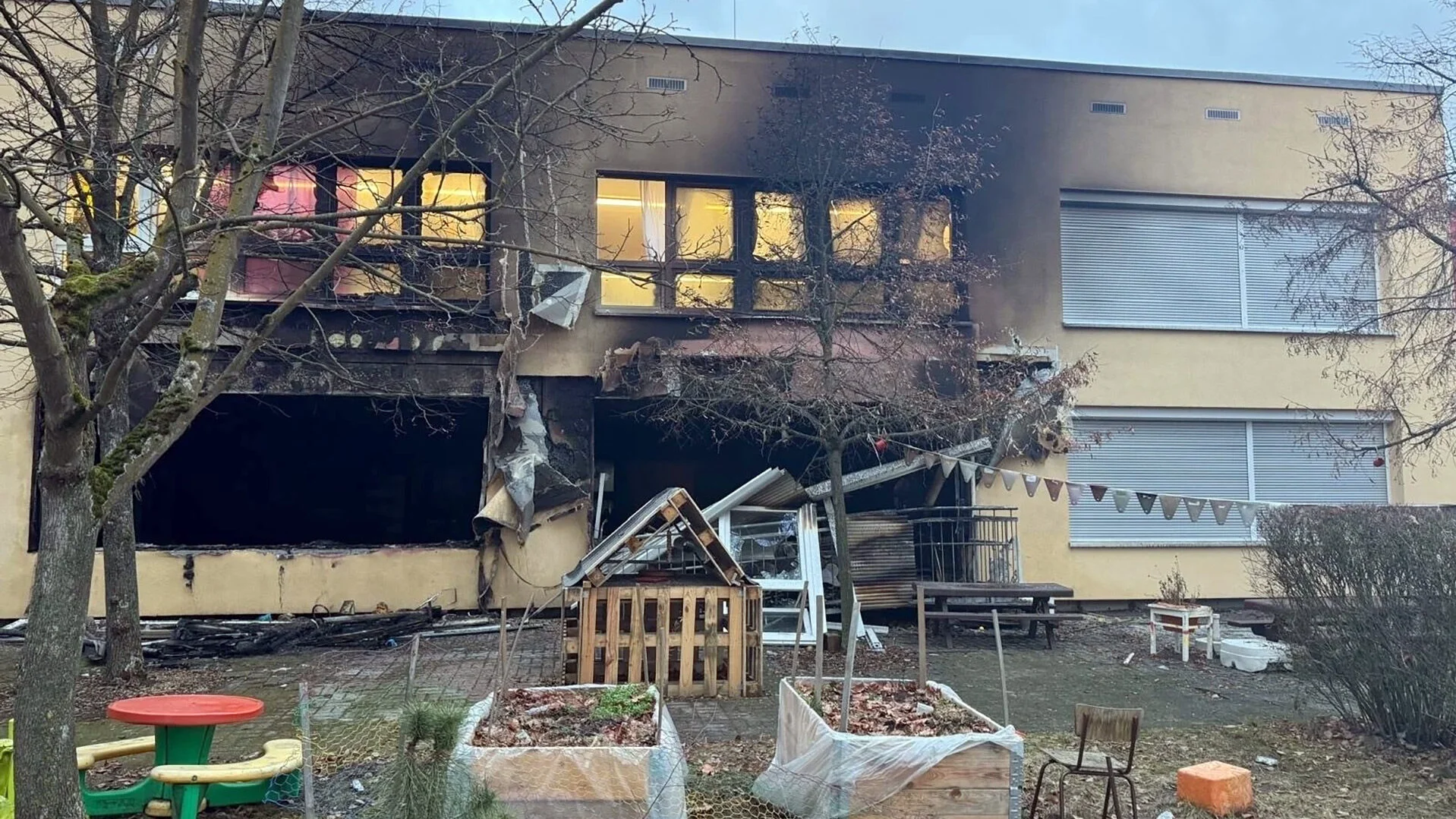 Auf dem Bild ist ein stark brandgeschädigtes Gebäude zu sehen. Es handelt sich um ein mehrstöckiges, beiges Gebäude, vermutlich eine Kita oder ein Kindergarten, das durch Feuer schwer beschädigt wurde. Erkennbare Details: Brandschäden: Der zentrale Bereich des Gebäudes zeigt massive Brandspuren mit geschwärzten Wänden und zerstörten Fenstern Außenbereich: Im Vordergrund ist ein Außenspielbereich mit typischen Kita-Elementen zu sehen, darunter ein kleiner grüner Tisch mit roter Platte, Hochbeete, ein Stuhl und verschiedene Spielgeräte Zerstörung: Die Fenster im Brandbereich sind ausgebrannt, es sind Trümmerteile sichtbar Winterliche Aufnahme: Kahle Bäume deuten auf eine kalte Jahreszeit hin
