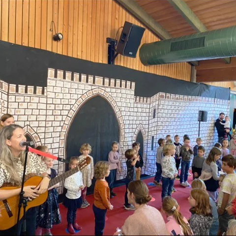 Das Foto zeigt eine Gruppe von Kindern, die auf einer Bühne stehen. Im Hintergrund ist eine Kulisse zu sehen, die wie eine mittelalterliche Burgmauer gestaltet ist. Eine Frau spielt Gitarre und begleitet die Kinder. Die Kinder stehen in einer Reihe und nehmen an einer Aufführung teil. Einige Erwachsene sind ebenfalls im Bild zu sehen. Die Szene findet in einem Raum mit Holzwänden und einer hohen Decke statt.