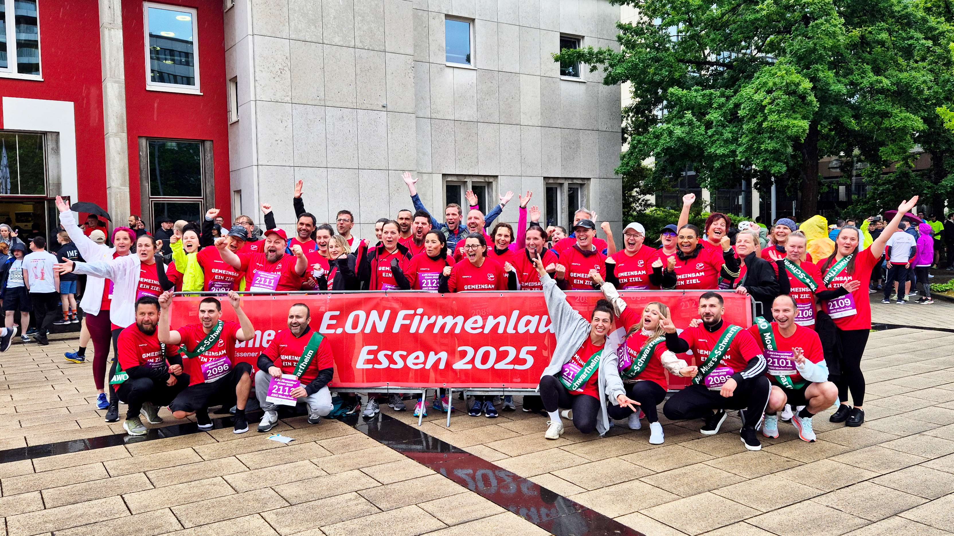 Auf dem Foto ist eine große Gruppe von Menschen zu sehen, die an einem Firmenlauf teilnimmt. Sie posieren gemeinsam für ein Gruppenfoto vor einem Gebäude. Viele von ihnen tragen rote T-Shirts mit der Aufschrift „E.ON Firmenlauf Essen 2025“, was auf ein Laufevent hinweist, das von der Firma E.ON organisiert wurde und im Jahr 2025 in Essen stattfindet. Die Stimmung ist ausgelassen und freudig – viele Personen lachen, heben die Arme oder zeigen Daumen nach oben. Vor der Gruppe wird ein großes rotes Banner mit der Aufschrift „E.ON Firmenlauf Essen 2025“ gehalten. Einige Teilnehmer tragen Startnummern an ihren T-Shirts. Im Hintergrund sind weitere Menschen zu sehen, die vermutlich auch Teil des Events sind, sowie ein modernes Gebäude mit roten und grauen Fassadenelementen. Das Wetter scheint leicht regnerisch zu sein, da der Boden nass ist. Zusammenfassend: Das Bild zeigt eine fröhliche und motivierte Laufgruppe beim E.ON-Firmenlauf 2025 in Essen.