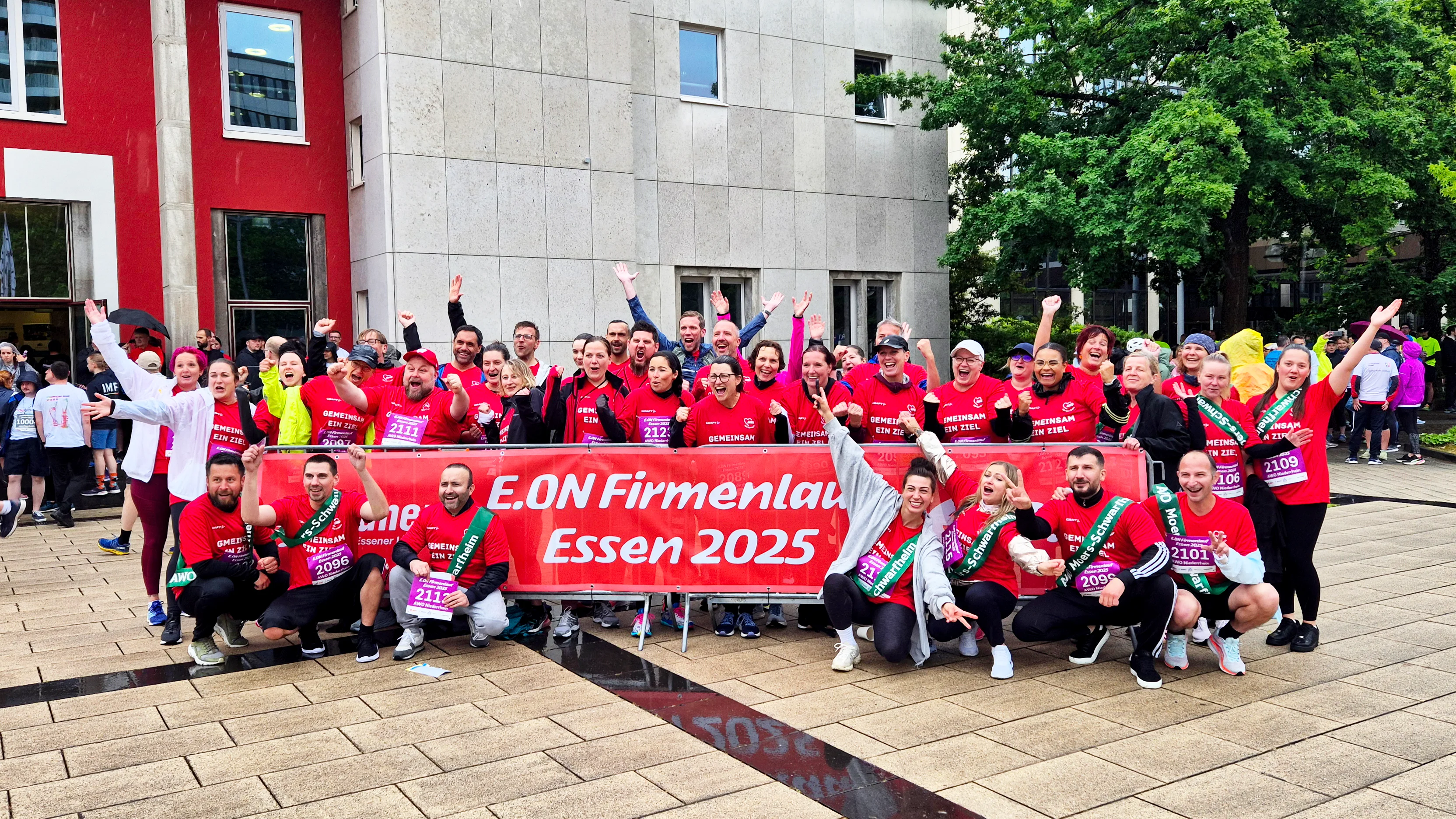 Auf dem Foto ist eine große Gruppe von Menschen zu sehen, die an einem Firmenlauf teilnimmt. Sie posieren gemeinsam für ein Gruppenfoto vor einem Gebäude. Viele von ihnen tragen rote T-Shirts mit der Aufschrift „E.ON Firmenlauf Essen 2025“, was auf ein Laufevent hinweist, das von der Firma E.ON organisiert wurde und im Jahr 2025 in Essen stattfindet. Die Stimmung ist ausgelassen und freudig – viele Personen lachen, heben die Arme oder zeigen Daumen nach oben. Vor der Gruppe wird ein großes rotes Banner mit der Aufschrift „E.ON Firmenlauf Essen 2025“ gehalten. Einige Teilnehmer tragen Startnummern an ihren T-Shirts. Im Hintergrund sind weitere Menschen zu sehen, die vermutlich auch Teil des Events sind, sowie ein modernes Gebäude mit roten und grauen Fassadenelementen. Das Wetter scheint leicht regnerisch zu sein, da der Boden nass ist. Zusammenfassend: Das Bild zeigt eine fröhliche und motivierte Laufgruppe beim E.ON-Firmenlauf 2025 in Essen.