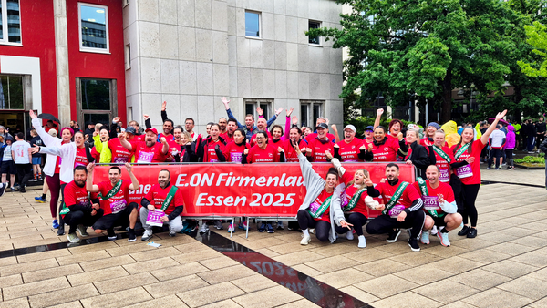 Auf dem Foto ist eine große Gruppe von Menschen zu sehen, die an einem Firmenlauf teilnimmt. Sie posieren gemeinsam für ein Gruppenfoto vor einem Gebäude. Viele von ihnen tragen rote T-Shirts mit der Aufschrift „E.ON Firmenlauf Essen 2025“, was auf ein Laufevent hinweist, das von der Firma E.ON organisiert wurde und im Jahr 2025 in Essen stattfindet. Die Stimmung ist ausgelassen und freudig – viele Personen lachen, heben die Arme oder zeigen Daumen nach oben. Vor der Gruppe wird ein großes rotes Banner mit der Aufschrift „E.ON Firmenlauf Essen 2025“ gehalten. Einige Teilnehmer tragen Startnummern an ihren T-Shirts. Im Hintergrund sind weitere Menschen zu sehen, die vermutlich auch Teil des Events sind, sowie ein modernes Gebäude mit roten und grauen Fassadenelementen. Das Wetter scheint leicht regnerisch zu sein, da der Boden nass ist. Zusammenfassend: Das Bild zeigt eine fröhliche und motivierte Laufgruppe beim E.ON-Firmenlauf 2025 in Essen.