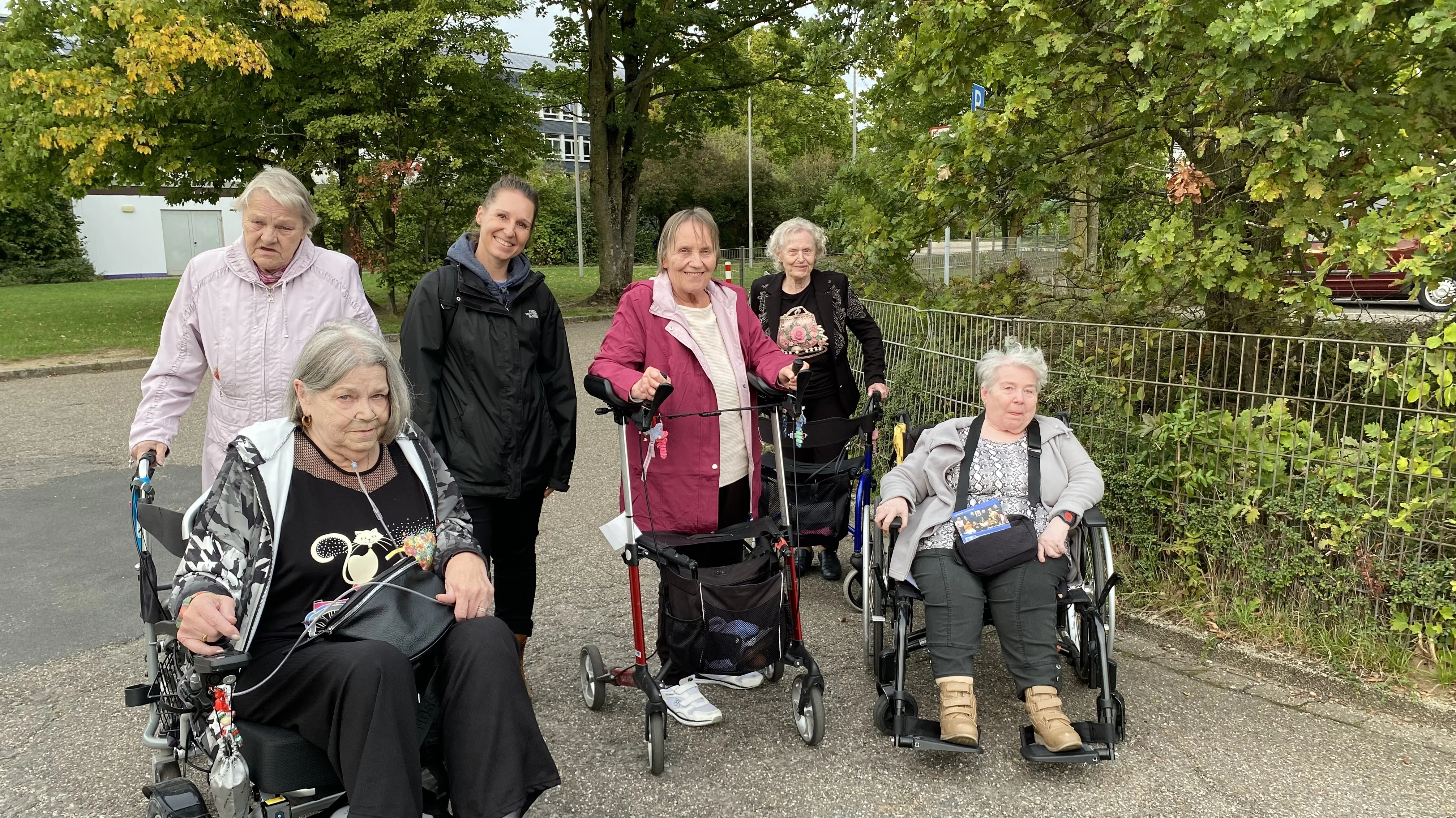 Auf dem Foto sind sechs Frauen zu sehen, die auf einer gepflasterten Fläche im Freien stehen oder sitzen. Im Vordergrund sind zwei Frauen in Rollstühlen zu sehen, und eine weitere Frau im Hintergrund benutzt einen Rollator. Eine jüngere Frau in einer schwarzen Jacke steht neben den anderen Frauen, und es gibt eine weitere Frau, die ebenfalls einen Rollator benutzt. Die Frauen scheinen in einer Gruppe unterwegs zu sein und stehen vor einer Kulisse aus Bäumen und Büschen.