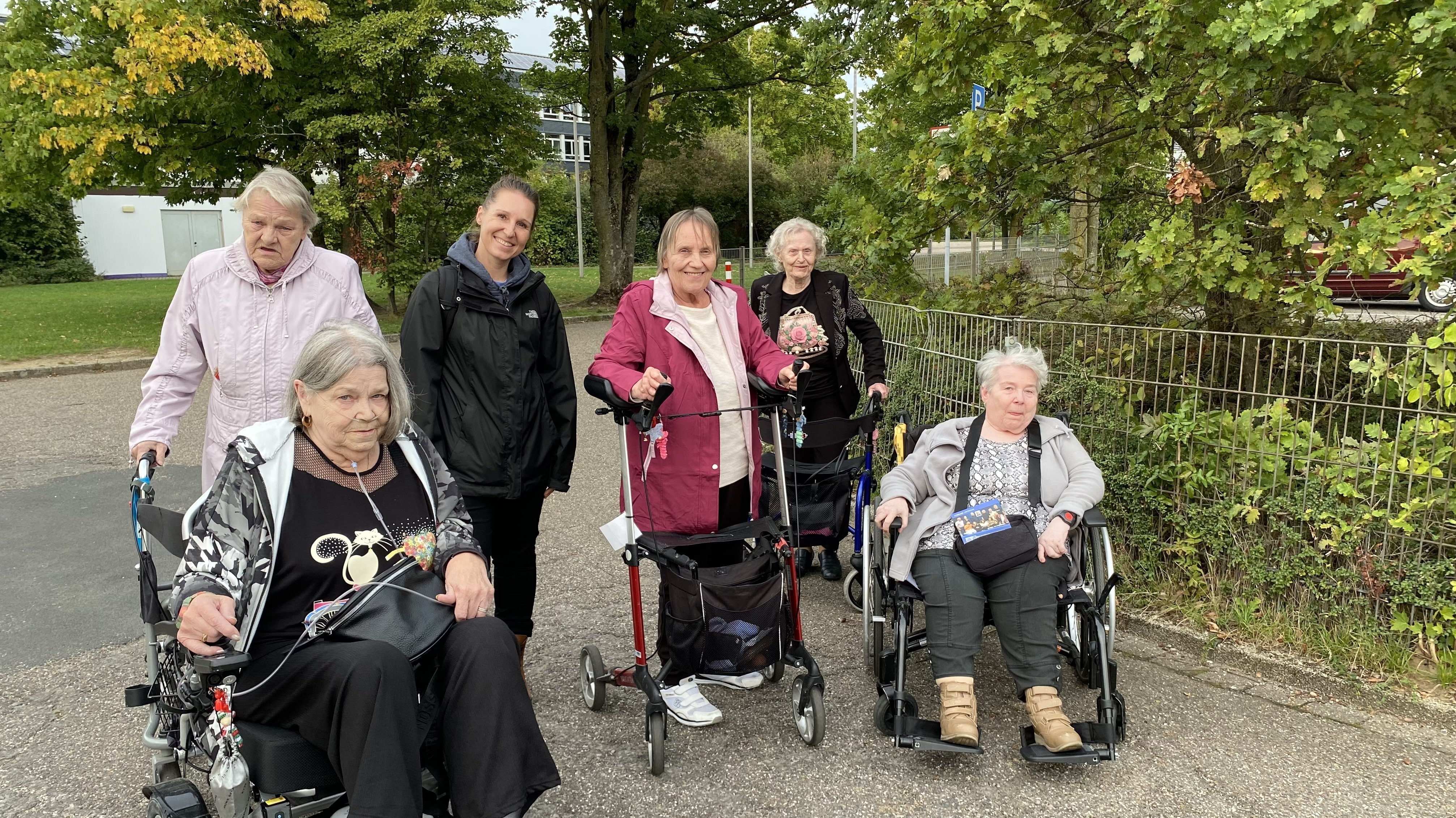 Auf dem Foto sind sechs Frauen zu sehen, die auf einer gepflasterten Fläche im Freien stehen oder sitzen. Im Vordergrund sind zwei Frauen in Rollstühlen zu sehen, und eine weitere Frau im Hintergrund benutzt einen Rollator. Eine jüngere Frau in einer schwarzen Jacke steht neben den anderen Frauen, und es gibt eine weitere Frau, die ebenfalls einen Rollator benutzt. Die Frauen scheinen in einer Gruppe unterwegs zu sein und stehen vor einer Kulisse aus Bäumen und Büschen.