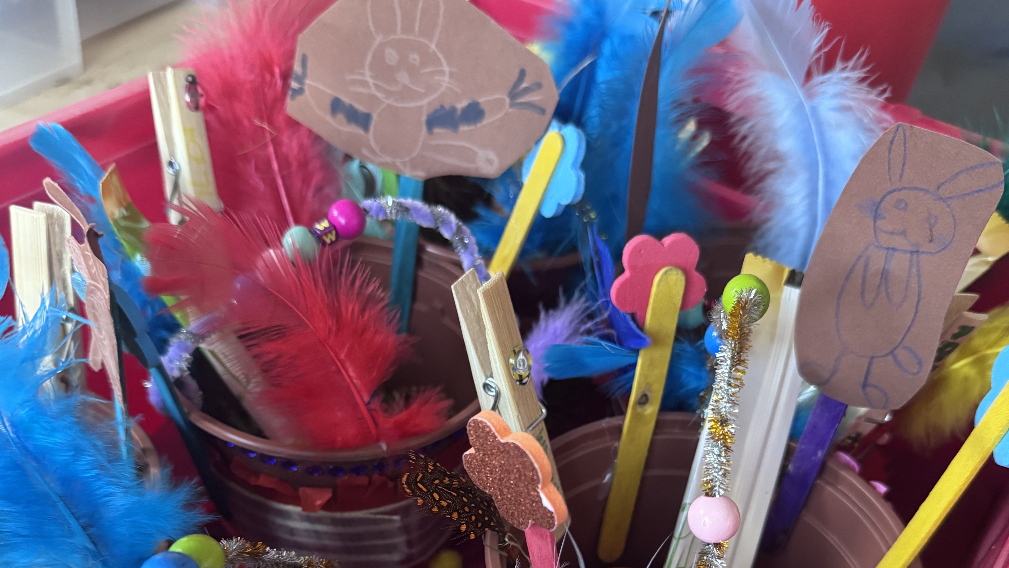 Auf dem Foto sind bunte, von Kindern gebastelte Oster- oder Frühlingskörbchen in kleinen Kunststoffbechern zu sehen. Die Becher sind mit vielen farbigen Federn, Pfeifenreinigern, Perlen, Holzklammern und aufgezeichneten Hasen verziert, und in den Bechern liegen kleine Süßigkeiten und ein gefärbtes Ei.