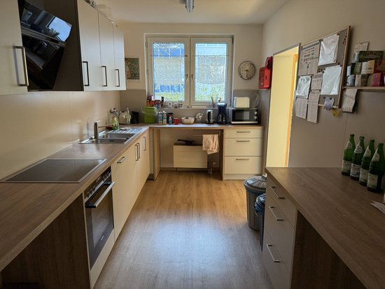 Das Bild zeigt eine moderne Küche mit Holzfußboden und weißen Schränken. Auf der linken Seite befinden sich ein Herd, ein Ofen und eine Spüle. In der Mitte des Raumes steht eine Arbeitsplatte mit verschiedenen Küchenutensilien, darunter ein Wasserkocher, eine Kaffeemaschine und Geschirr. Rechts an der Wand hängen verschiedene Notizen und Papiere. Es gibt auch mehrere grüne Glasflaschen auf einer Arbeitsfläche rechts im Vordergrund. Am Ende des Raumes sind zwei Fenster, durch die Tageslicht hereinfällt.