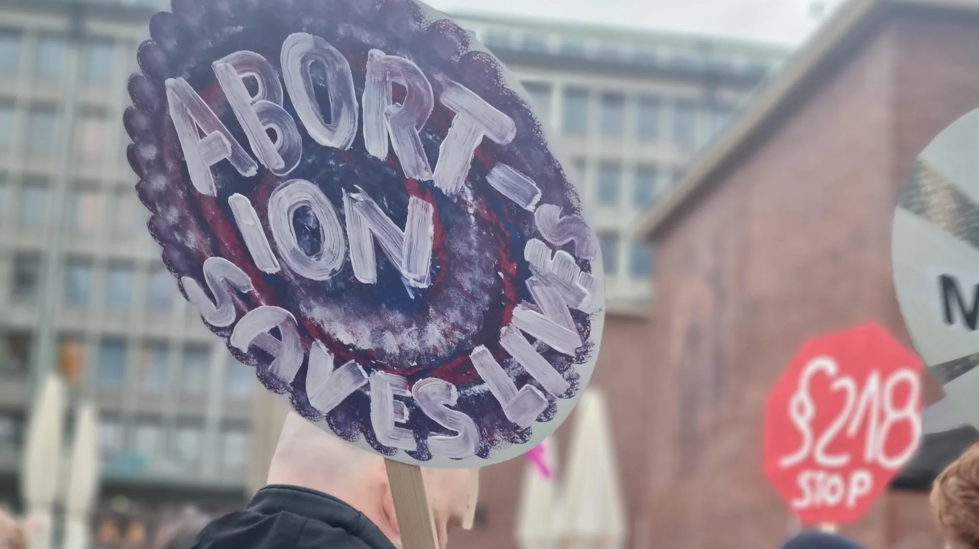 Auf dem Foto ist eine Demonstration oder eine ähnliche Versammlung zu sehen. Im Vordergrund hält eine Person, von hinten aufgenommen, ein selbstgemaltes, rundes Schild, auf dem in weißen und lilafarbenen Buchstaben steht: "ABORTION SAVES LIVES" (Abtreibung rettet Leben). Im Hintergrund sind weitere Menschen und einige verschwommen Gebäude erkennbar, sowie ein weiteres rotes, sechseckiges Schild mit der Aufschrift "§218 STOP".