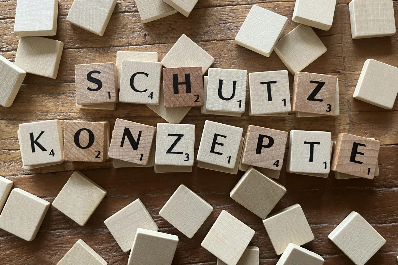 Auf dem Foto sind Holzplättchen zu sehen, die ähnlich wie bei dem Spiel Scrabble verwendet werden. Die Plättchen sind so angeordnet, dass sie die Wörter “SCHUTZ” und “KONZEPTE” bilden.