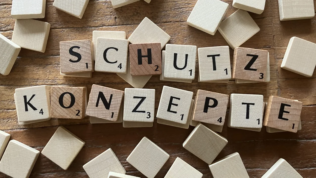 Auf dem Foto sind Scrabble-ähnliche Holzbuchstabensteine zu sehen, die auf einem hölzernen Untergrund verteilt sind. Einige der Steine sind zu den Wörtern „SCHUTZ KONZEPTE“ zusammengesetzt, während andere verstreut herumliegen.