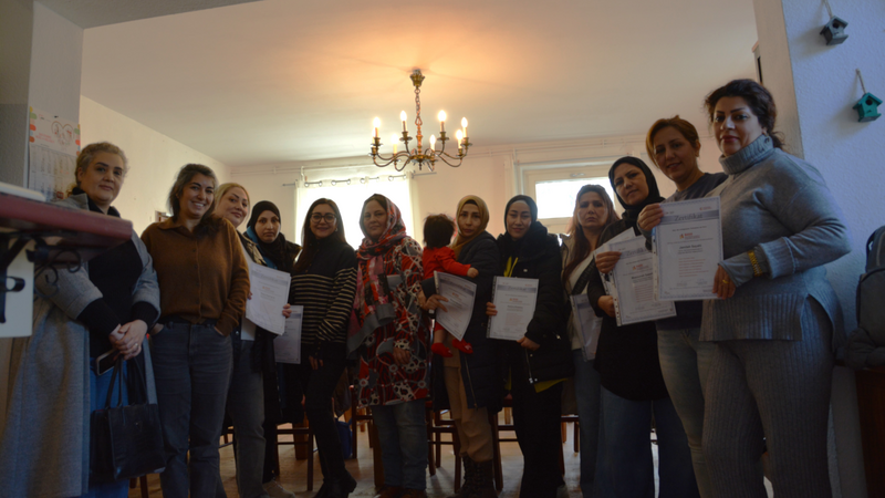 Auf dem Foto ist eine Gruppe von etwa 12-13 Frauen zu sehen, die in einem hellen Raum zusammenstehen und eine Art Abschlussfeier oder Zertifikatsverleihung feiern. Viele der Frauen halten Urkunden oder Zertifikate in den Händen und lächeln in die Kamera. Die Gruppe ist vielfältig - einige Frauen tragen Kopftücher (Hijab), andere nicht. Sie stehen in einer Reihe nebeneinander. Im Zentrum der Gruppe ist eine Frau in einem auffälligen rot-gemusterten Kleid besonders zu erkennen. Die Kleidung reicht von lässig (Jeans und Pullover) bis förmlicher. Der Raum wirkt wie ein Gemeinschaftsraum oder Schulungsraum mit weißen Wänden, einem Kronleuchter an der Decke, Fenstern im Hintergrund und dekorativen Vogelhäuschen an der Wand. Im Vordergrund sind Stühle zu sehen, was darauf hindeutet, dass zuvor eine Veranstaltung oder ein Kurs stattgefunden hat. Die Atmosphäre ist fröhlich und feierlich - es scheint ein erfolgreicher Abschluss eines Kurses oder Programms zu sein, vermutlich im Kontext von Bildung oder Integration.