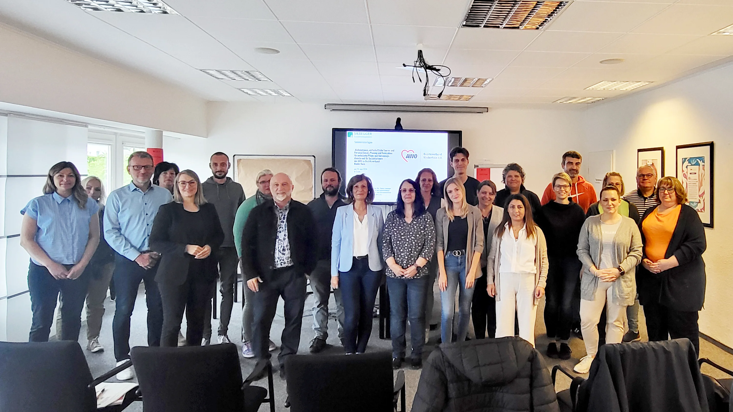 Das Foto zeigt eine Gruppe von Personen in einem Raum, der für ein Meeting oder eine Präsentation eingerichtet ist. Es gibt Reihen von Stühlen, die auf eine Leinwand vorne ausgerichtet sind. Auf der Leinwand sind Text und Grafiken zu sehen, die von dieser Perspektive aus jedoch nicht lesbar sind. Der Raum verfügt über Deckenleuchten und ein Fenster mit teilweise heruntergelassenen Jalousien. Die Personen sind unterschiedlich gekleidet, was auf eine formelle oder geschäftlich-legere Umgebung hindeutet. 