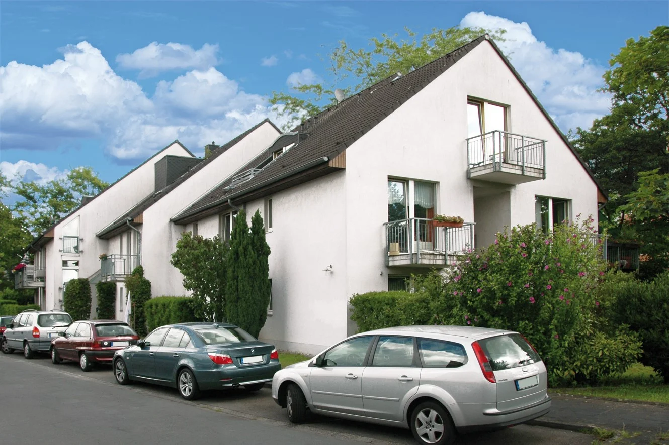 Auf dem Foto ist eine Reihenhauszeile zu sehen, die in der Mitte eine Art Giebel oder einen höheren Gebäudeteil hat. Das Gebäude ist weiß gestrichen und hat ein dunkles Dach. Es gibt Balkone mit Metallgeländern und große Fenster. Vor dem Haus sind mehrere Autos am Straßenrand geparkt, darunter ein silberner Kombi, eine dunkelgrüne Limousine und eine bordeauxrote Limousine. Es gibt auch etwas Grünfläche, wie Hecken und Bäume, die das Gebäude umgeben. Der Himmel ist blau mit einigen weißen Wolken.