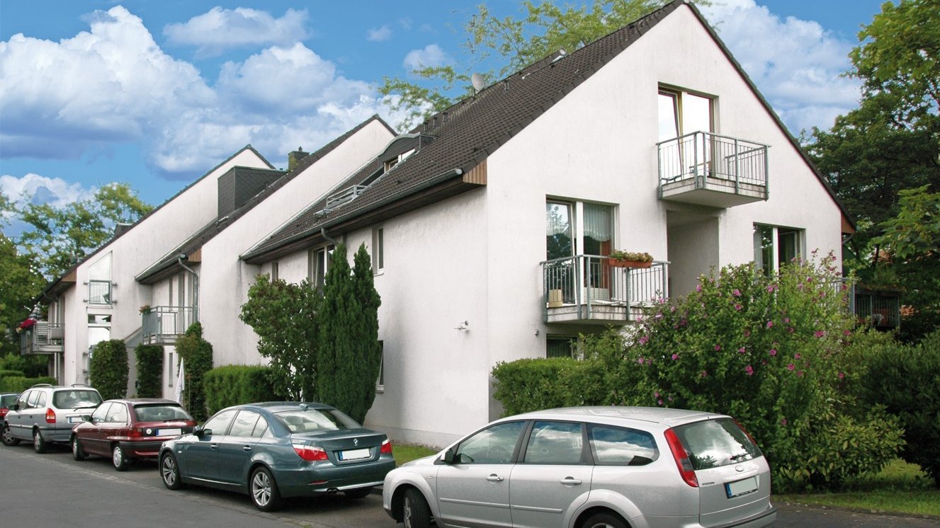  Auf dem Foto ist eine Reihenhauszeile zu sehen, die in der Mitte eine Art Giebel oder einen höheren Gebäudeteil hat. Das Gebäude ist weiß gestrichen und hat ein dunkles Dach. Es gibt Balkone mit Metallgeländern und große Fenster. Vor dem Haus sind mehrere Autos am Straßenrand geparkt, darunter ein silberner Kombi, eine dunkelgrüne Limousine und eine bordeauxrote Limousine. Es gibt auch etwas Grünfläche, wie Hecken und Bäume, die das Gebäude umgeben. Der Himmel ist blau mit einigen weißen Wolken.