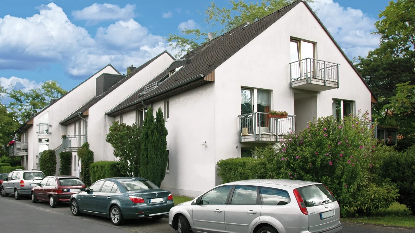  Auf dem Foto ist eine Reihenhauszeile zu sehen, die in der Mitte eine Art Giebel oder einen höheren Gebäudeteil hat. Das Gebäude ist weiß gestrichen und hat ein dunkles Dach. Es gibt Balkone mit Metallgeländern und große Fenster. Vor dem Haus sind mehrere Autos am Straßenrand geparkt, darunter ein silberner Kombi, eine dunkelgrüne Limousine und eine bordeauxrote Limousine. Es gibt auch etwas Grünfläche, wie Hecken und Bäume, die das Gebäude umgeben. Der Himmel ist blau mit einigen weißen Wolken.