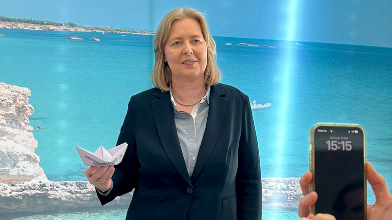 Auf dem Foto ist eine Frau in einem dunklen Blazer und einer hellgrauen Bluse zu sehen, die ein gefaltetes Papierboot in ihrer linken Hand hält. Sie steht vor einem Hintergrund, der eine Küstenlandschaft mit blauem Meer, hellem Sand und Felsen zeigt. Rechts im Bild ist ein Teil einer Person zu sehen, die ein Smartphone in beiden Händen hält und die Frau zu fotografieren scheint. Auf dem Display des Smartphones ist die Uhrzeit 15:15 zu erkennen. Links im Bild ist ein runder Tisch mit einigen Papierdokumenten darauf angeschnitten.