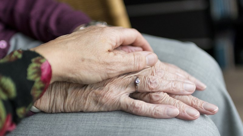 Auf dem Foto sieht man die gefalteten Hände einer älteren Person, auf denen behutsam die Hand einer anderen Person liegt. Die Szene vermittelt Nähe, Fürsorge und Unterstützung, etwa im Kontext von Pflege, Begleitung oder Trost.