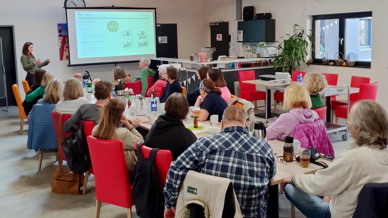 Auf dem Bild sieht man eine Gruppenveranstaltung in einem hellen Gemeinschaftsraum. Die Teilnehmenden sitzen an mehreren Tischen mit roten Stühlen, trinken und sprechen miteinander, während vorne eine Person neben einer Leinwand steht und eine Präsentation hält.