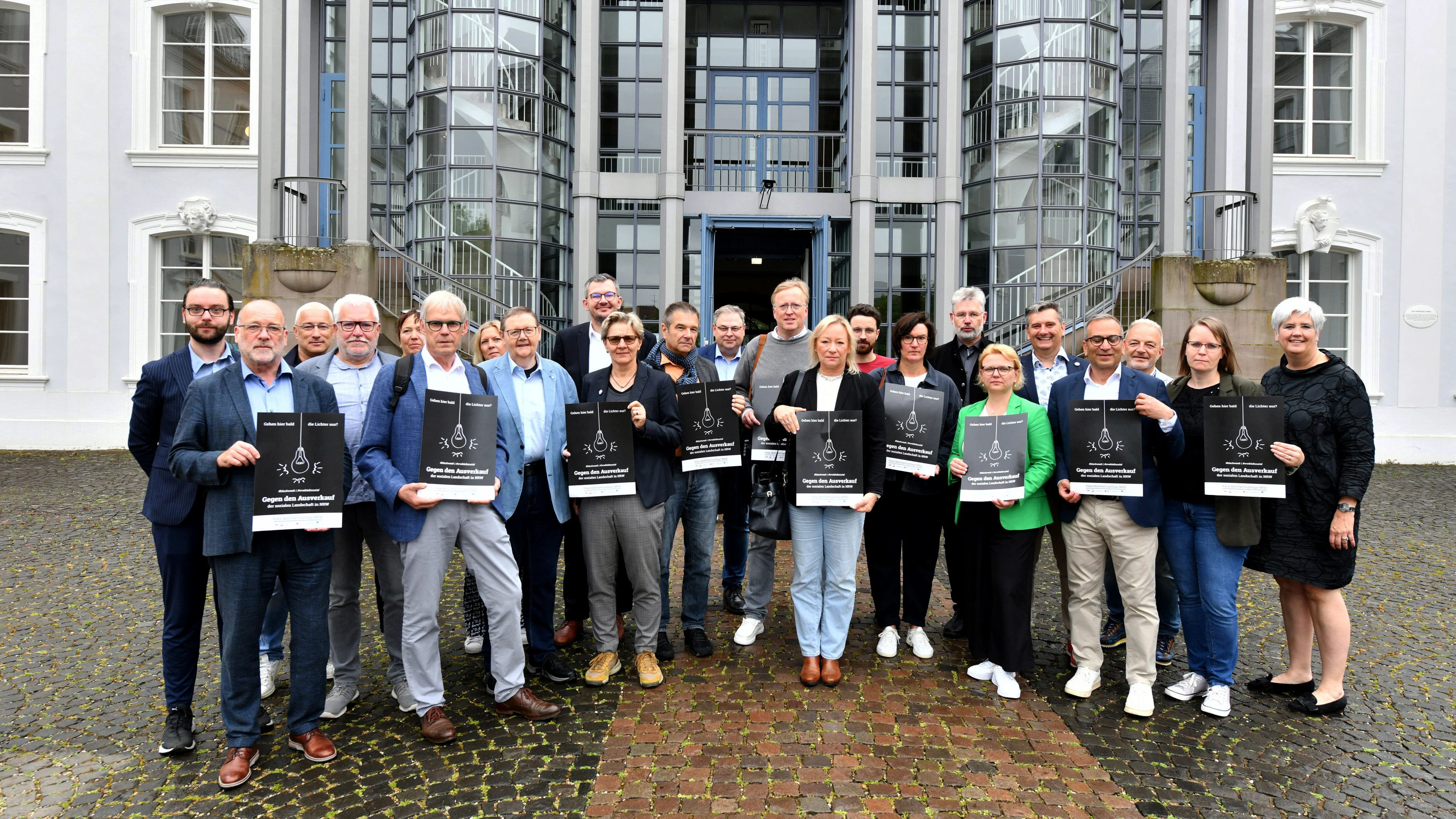 Das Foto zeigt eine Gruppe von Personen vor einem Gebäude mit modernen Glastüren und traditioneller Architektur. Einzelne Person halten das Kampagnenplakat zur #blackweek hoch auf dem steht: Gehen hier bald die Lichter aus? Gegen den Ausverkauf der sozialen Landschaft in NRW. 