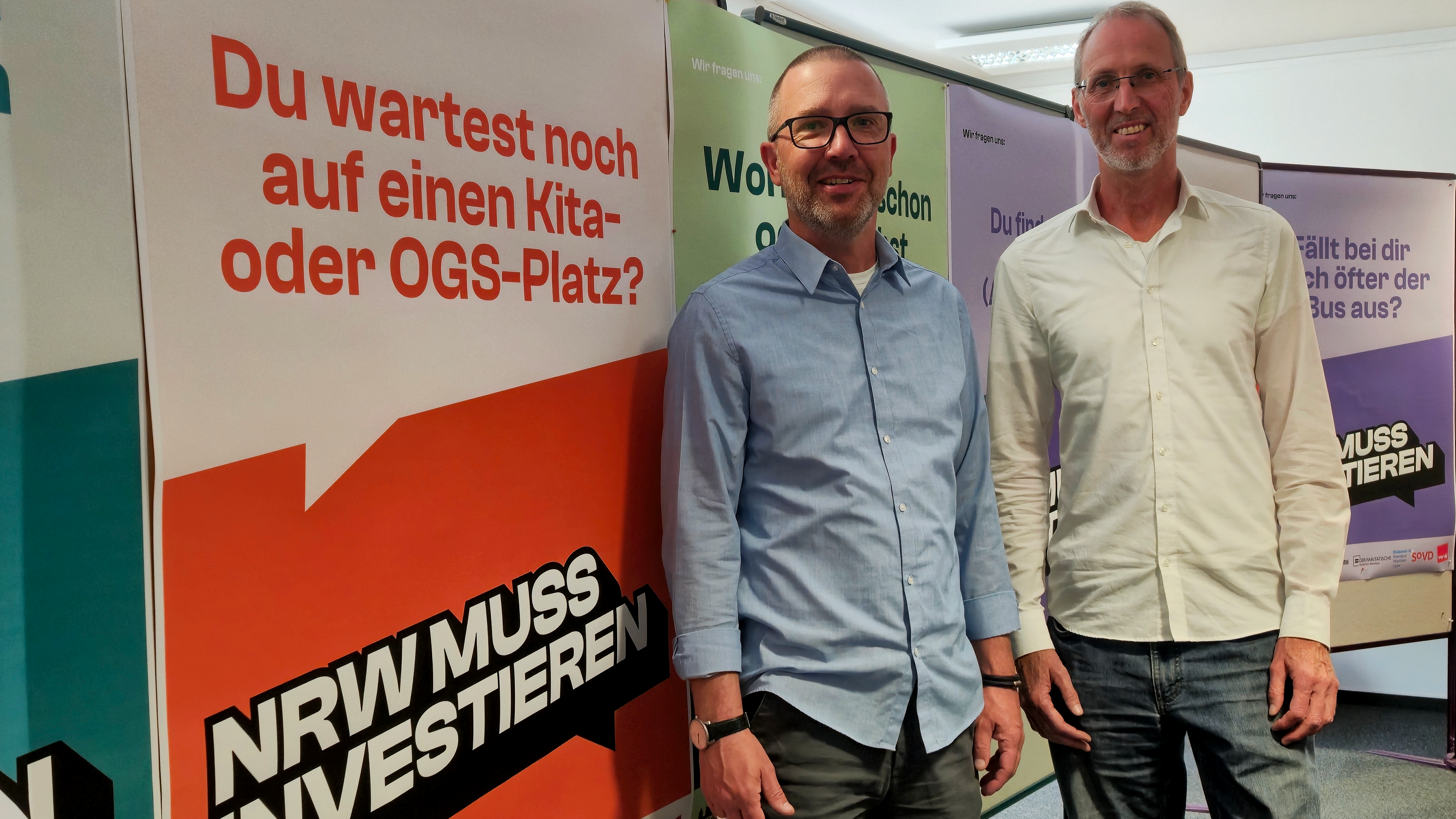 Auf dem Foto sind zwei Männer zu sehen, die nebeneinander stehen und in die Kamera lächeln. Sie stehen vor mehreren bunten Plakaten mit politischen oder gesellschaftlichen Botschaften auf Deutsch. Eines der Plakate lautet: „Du wartest noch auf einen Kita- oder OGS-Platz?“ Darunter steht in fetter Schrift: „NRW MUSS INVESTIEREN“ Weitere Plakate im Hintergrund tragen ähnliche Fragen wie: „Worauf wartest du schon ganz lange?“ „Du findest keinen (Ausbildungs-)Platz?“ „Fällt bei dir auch öfter der Bus aus?“ Am unteren Rand der Plakate befinden sich Logos, u.a. vom „Sozialverband Deutschland (SoVD)“. Das Bild scheint im Kontext einer Kampagne oder Pressekonferenz aufgenommen worden zu sein, die auf Missstände in Nordrhein-Westfalen (NRW) hinweist und Investitionen fordert – insbesondere in Bildung, Infrastruktur und soziale Angebote.