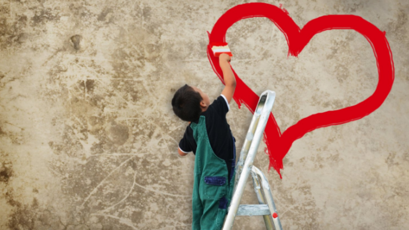 Auf dem Bild steht ein Kind auf einer kleinen Leiter und malt mit einem Pinsel ein großes rotes Herz auf eine leicht bröckelige, beige Wand.