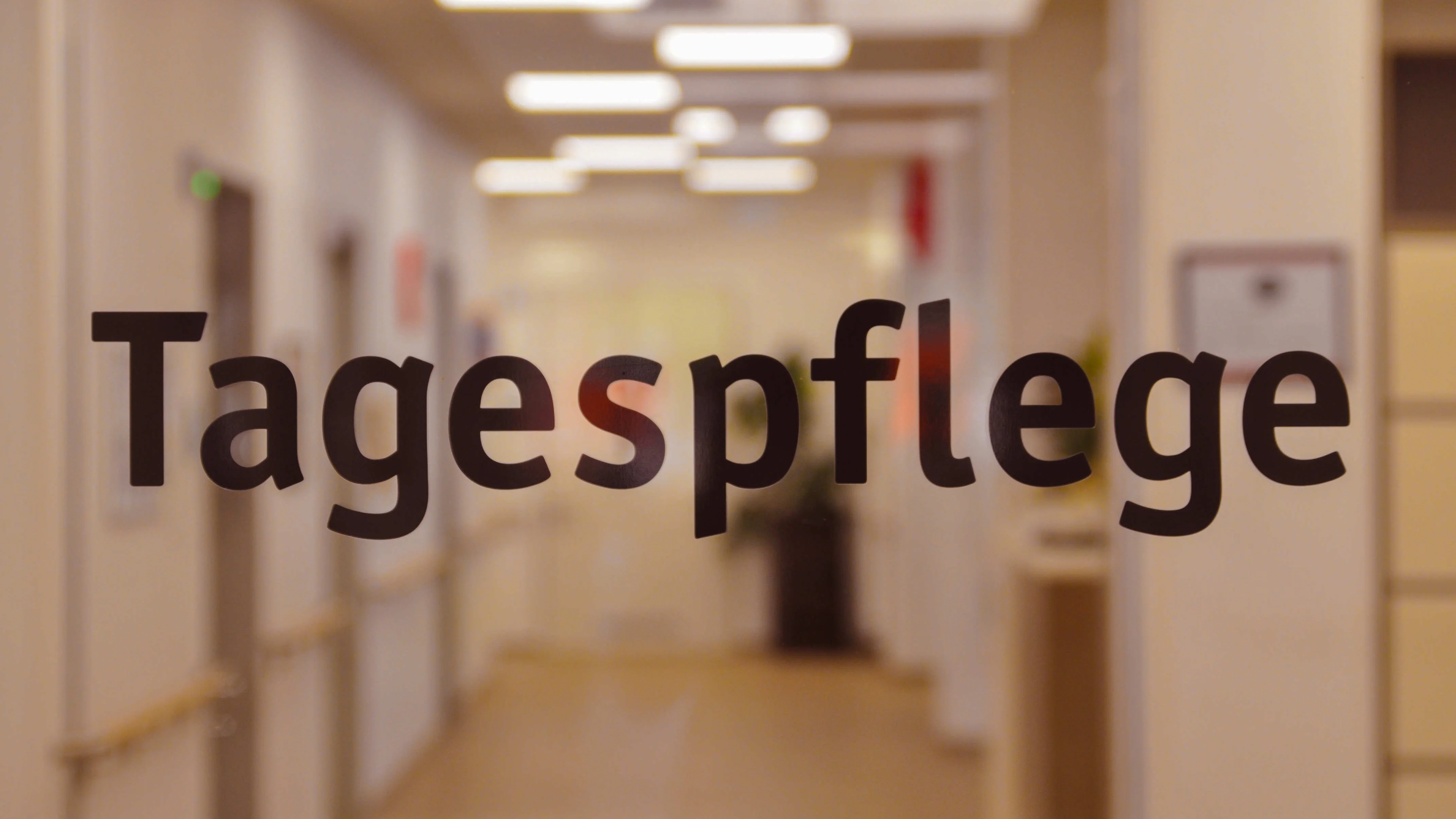 Zu sehen ist ein unscharf fotografierter, hell beleuchteter Flur einer Einrichtung. Im Vordergrund steht auf einer Glastür oder Glasscheibe groß der Schriftzug „Tagespflege“. Im Hintergrund erkennt man unter anderem Deckenleuchten, Türen, Handläufe und einen Flurbereich.