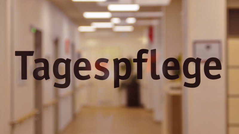 Zu sehen ist ein unscharf fotografierter, hell beleuchteter Flur einer Einrichtung. Im Vordergrund steht auf einer Glastür oder Glasscheibe groß der Schriftzug „Tagespflege“. Im Hintergrund erkennt man unter anderem Deckenleuchten, Türen, Handläufe und einen Flurbereich.