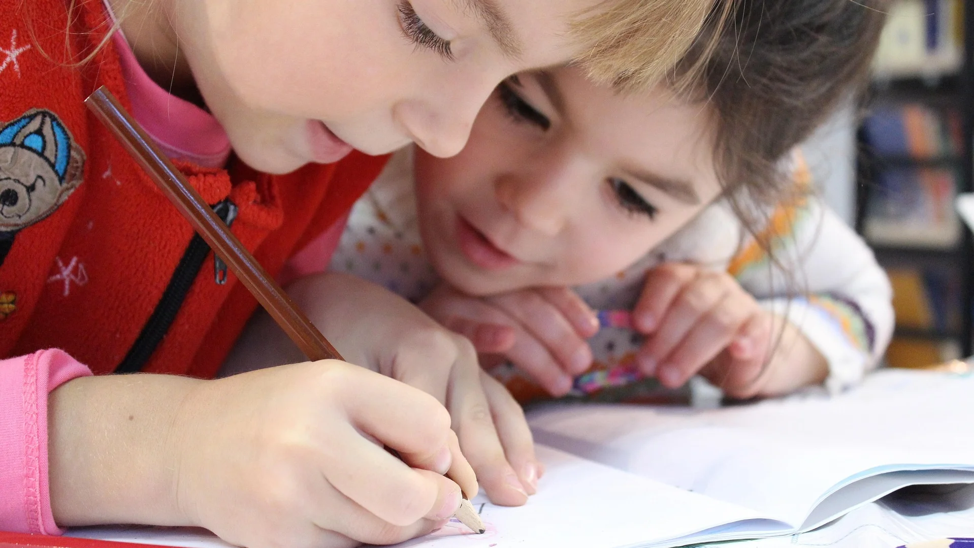 Auf dem Bild sind zwei junge Kinder zu sehen, die eng beieinander über einem geöffneten Heft oder Buch sitzen. Ein Kind, das eine rote Jacke oder einen Pullover trägt, hält einen Bleistift in der Hand und zeichnet oder schreibt in das Heft. Das andere Kind lehnt sich von hinten leicht nach vorne, um ebenfalls auf das Heft zu schauen. Im Vordergrund liegen noch ein paar weitere Buntstifte, unter anderem ein roter und ein blauer. Der Hintergrund ist unscharf, scheint aber Bücherregale oder eine Bibliothek zu zeigen, was auf eine Lern- oder Leseumgebung hindeutet.