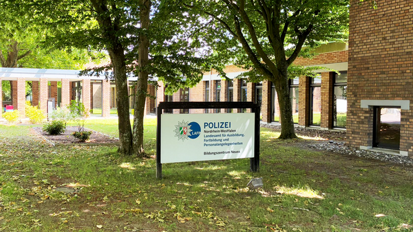 Auf dem Foto ist ein Gebäude mit einem Schild im Vordergrund zu sehen, das teilweise von Bäumen beschattet wird. Das Schild trägt die Aufschrift: POLIZEI Nordrhein-Westfalen Landesamt für Ausbildung, Fortbildung und Personalangelegenheiten der Polizei NRW (LAFP NRW) Bildungszentrum Neuss Es handelt sich also um ein Ausbildungs- und Fortbildungszentrum der Polizei Nordrhein-Westfalen in der Stadt Neuss. Das Gelände ist gepflegt, mit Rasen, Bäumen und einem modernen Backsteingebäude im Hintergrund. Links im Bild sind auch ein paar Fahrräder zu sehen.